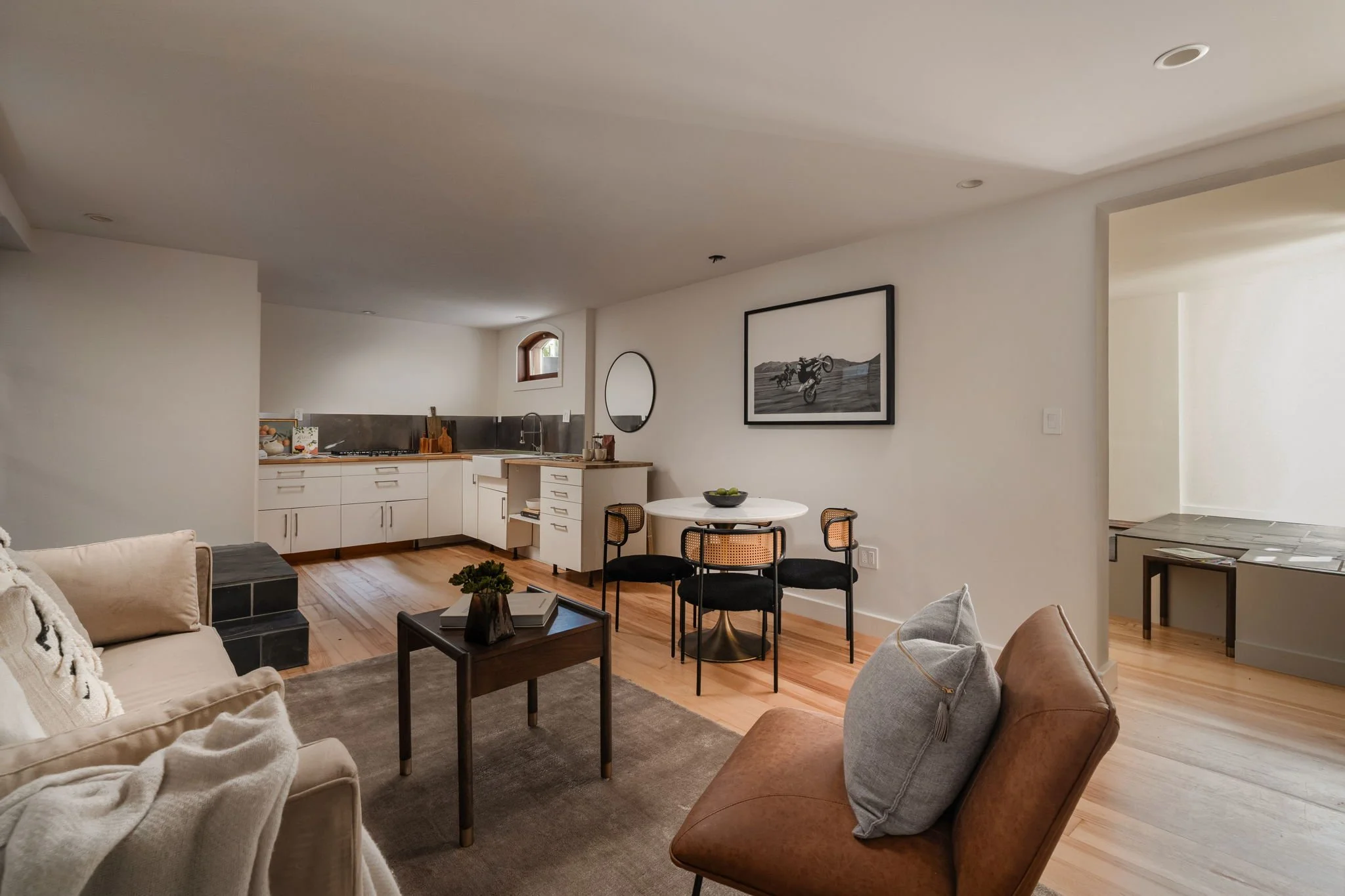 Living room with beige and brown furniture, a sofa, chairs, a coffee table with magazines, a dining table with chairs, and wall art, with a kitchen area and small window