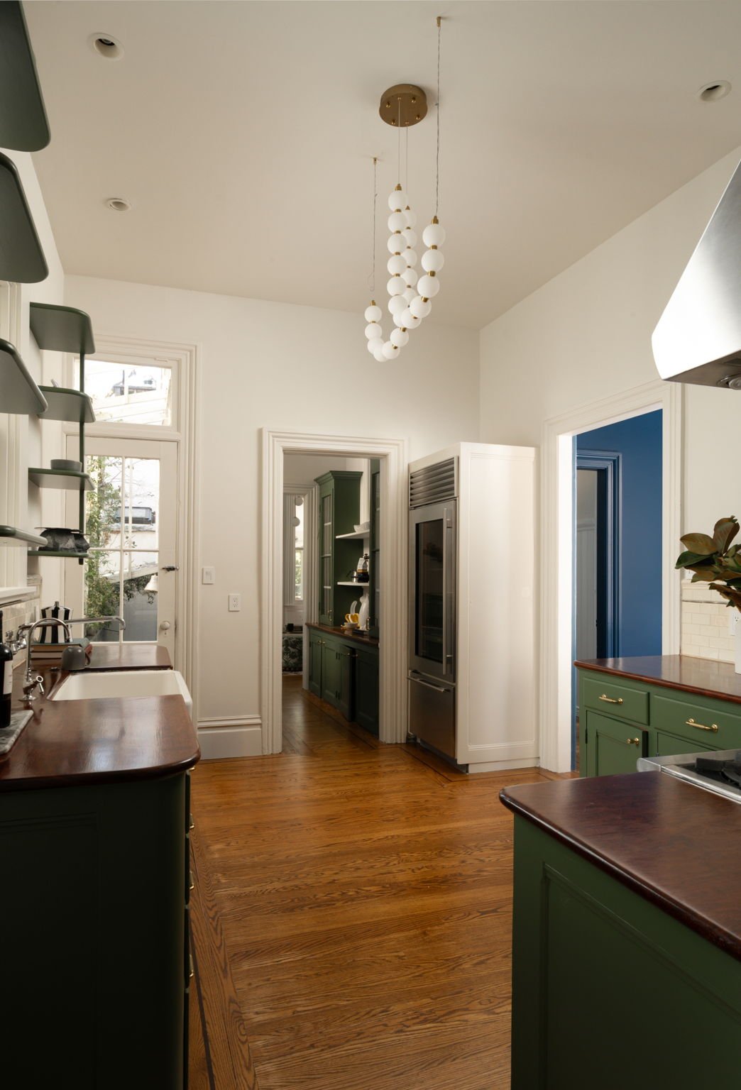 A kitchen with white walls, green cabinets, wooden countertops, hardwood flooring, a modern hanging light fixture with white globes, stainless steel appliances, and a door leading outside.