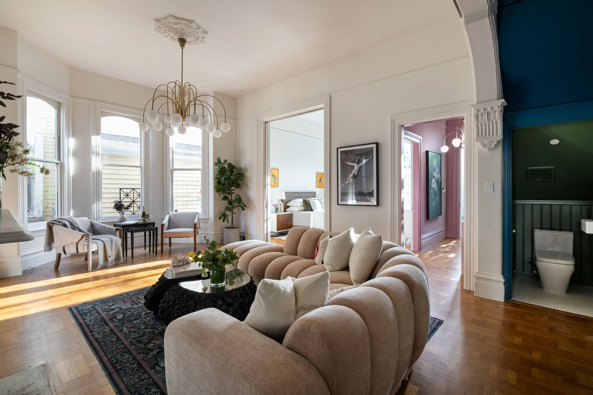 Living room with beige sectional sofa, armchair, patterned rug, and a black coffee table with a vase of greenery, bright windows, and artwork on the walls.