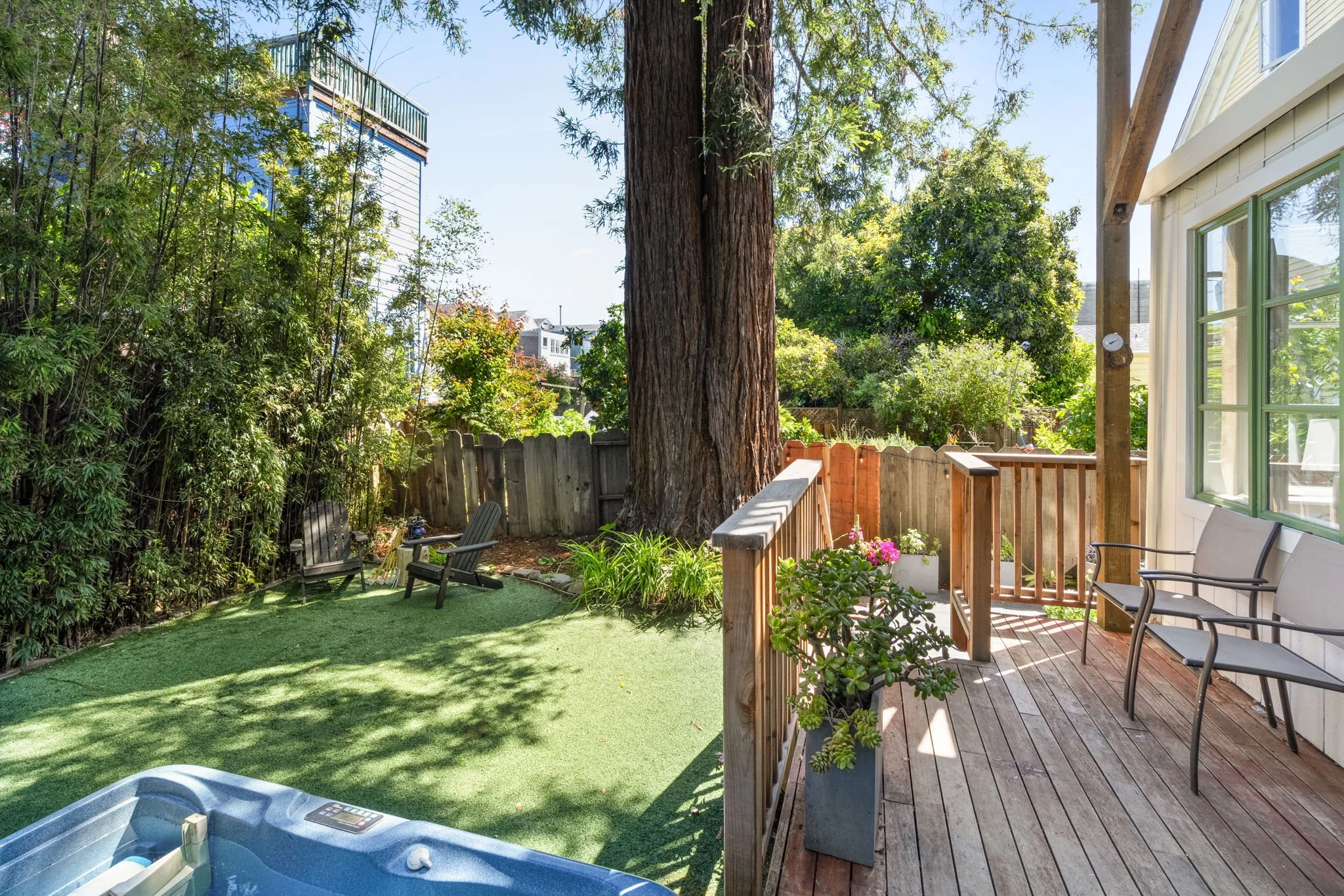 A backyard with a wooden deck and patio chairs, a green artificial lawn, various trees and shrubs, and a large tree trunk in the center. The sky is clear and blue.