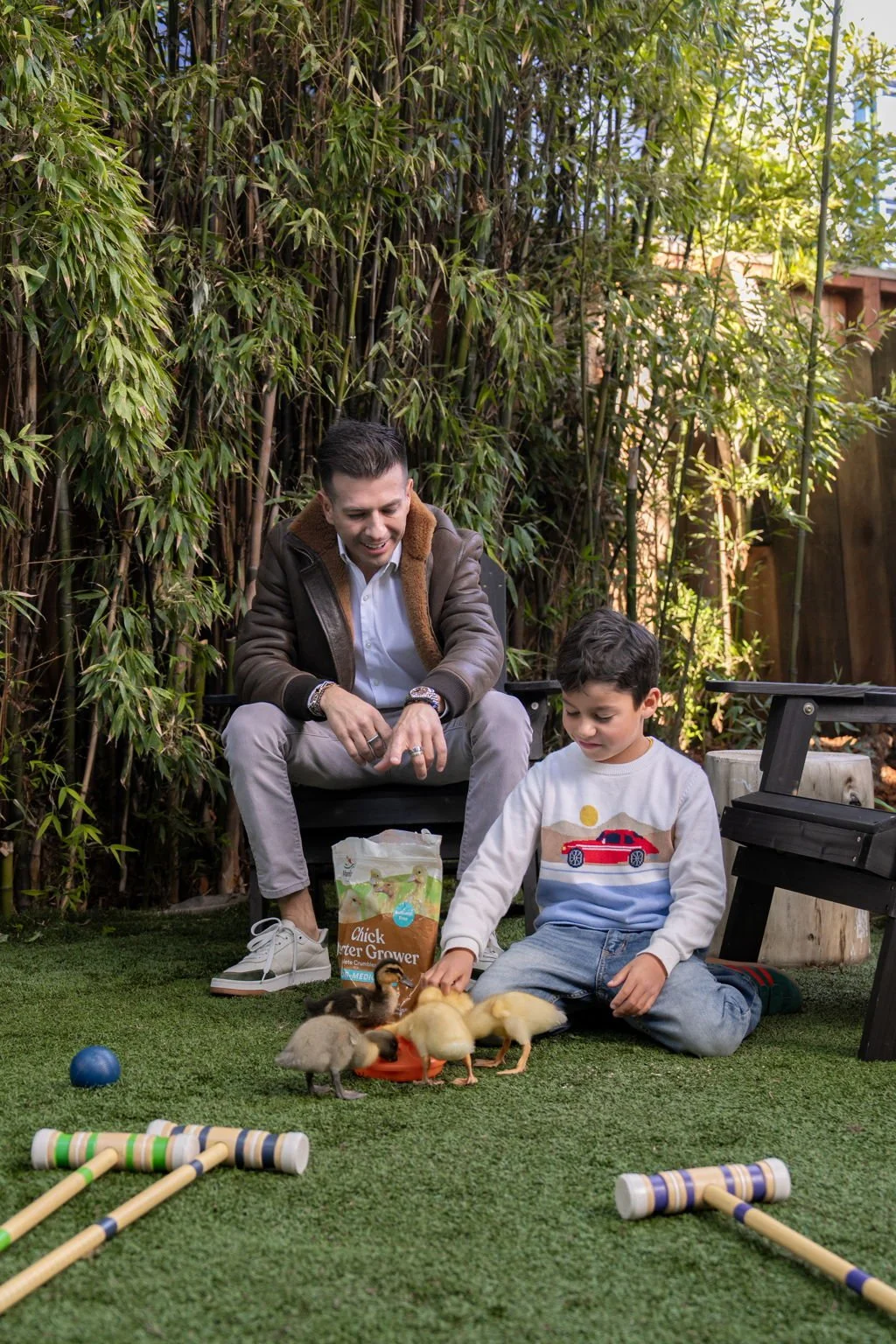 A man and a boy sitting on grass in a backyard, feeding a group of ducklings. The man is smiling and watching the ducklings, while the boy is focused on feeding them. There is a bag of chicken starter grower feed near them, and some outdoor toys arou