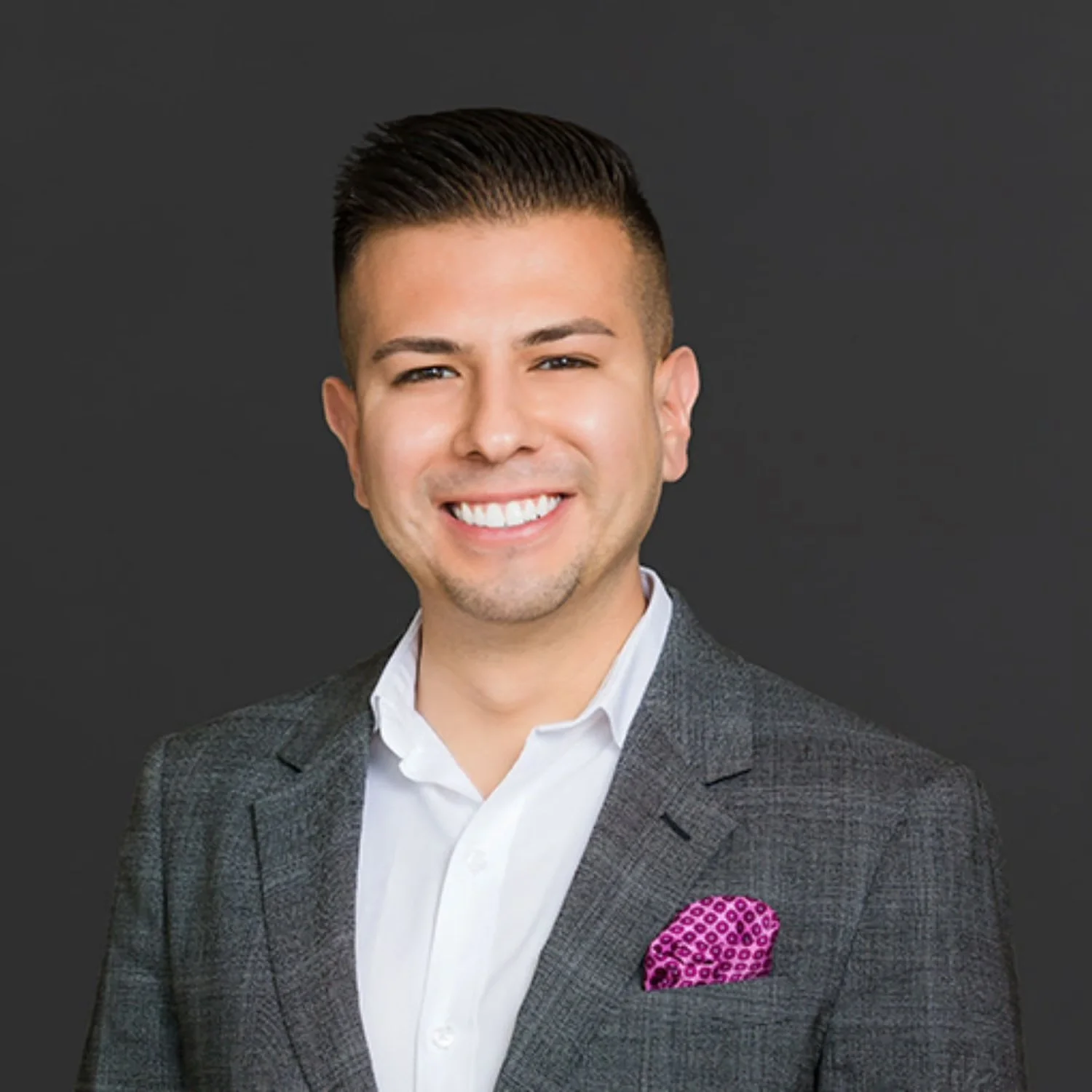 A young man smiling, wearing a white shirt, gray checked blazer, and a pink pocket square, standing against a dark gray background.