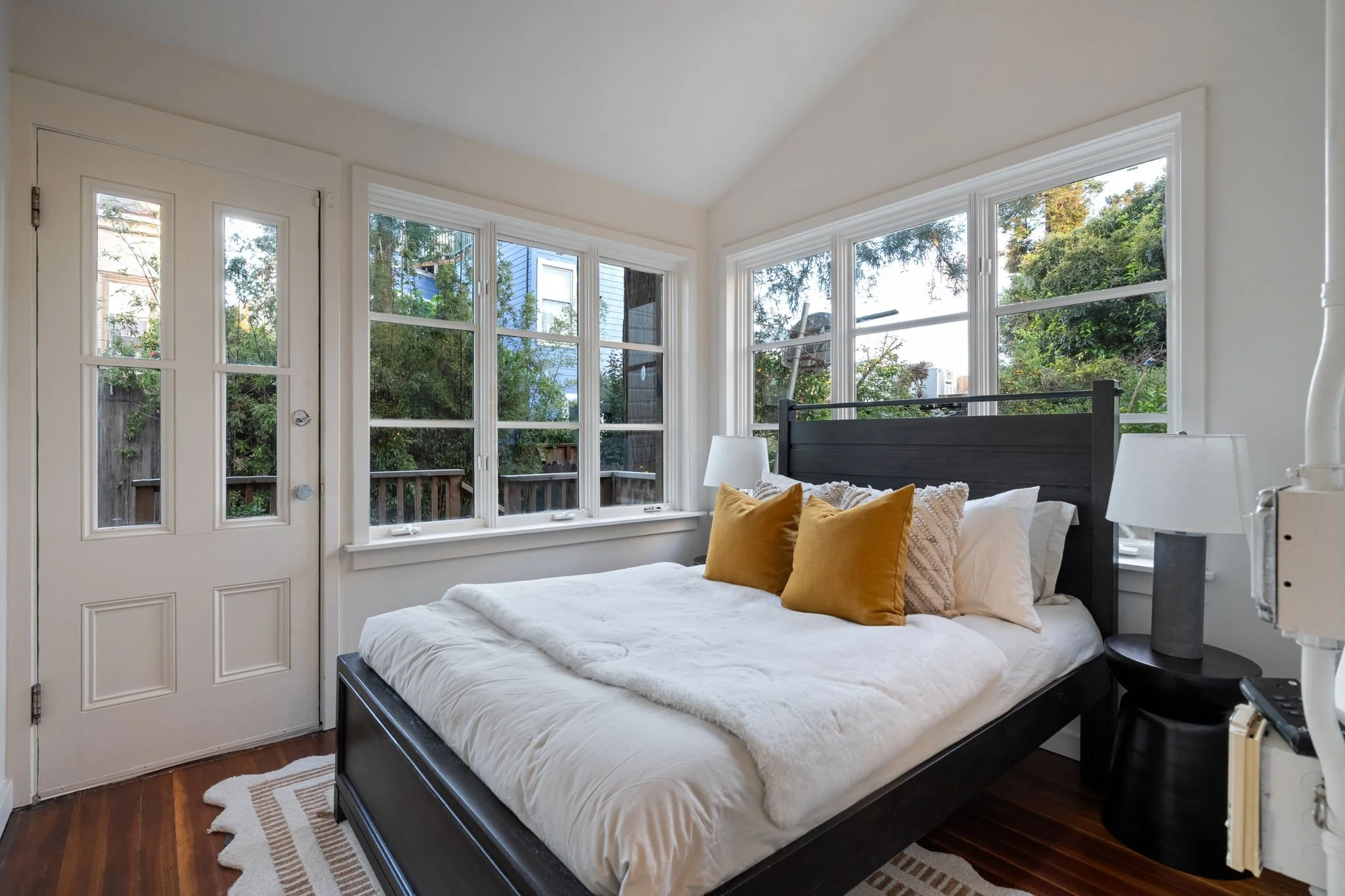 A bedroom with large windows showing trees outside, a bed with white linens and mustard-colored pillows, a black headboard, a small bedside table with a white lamp, and a door with four glass panes.