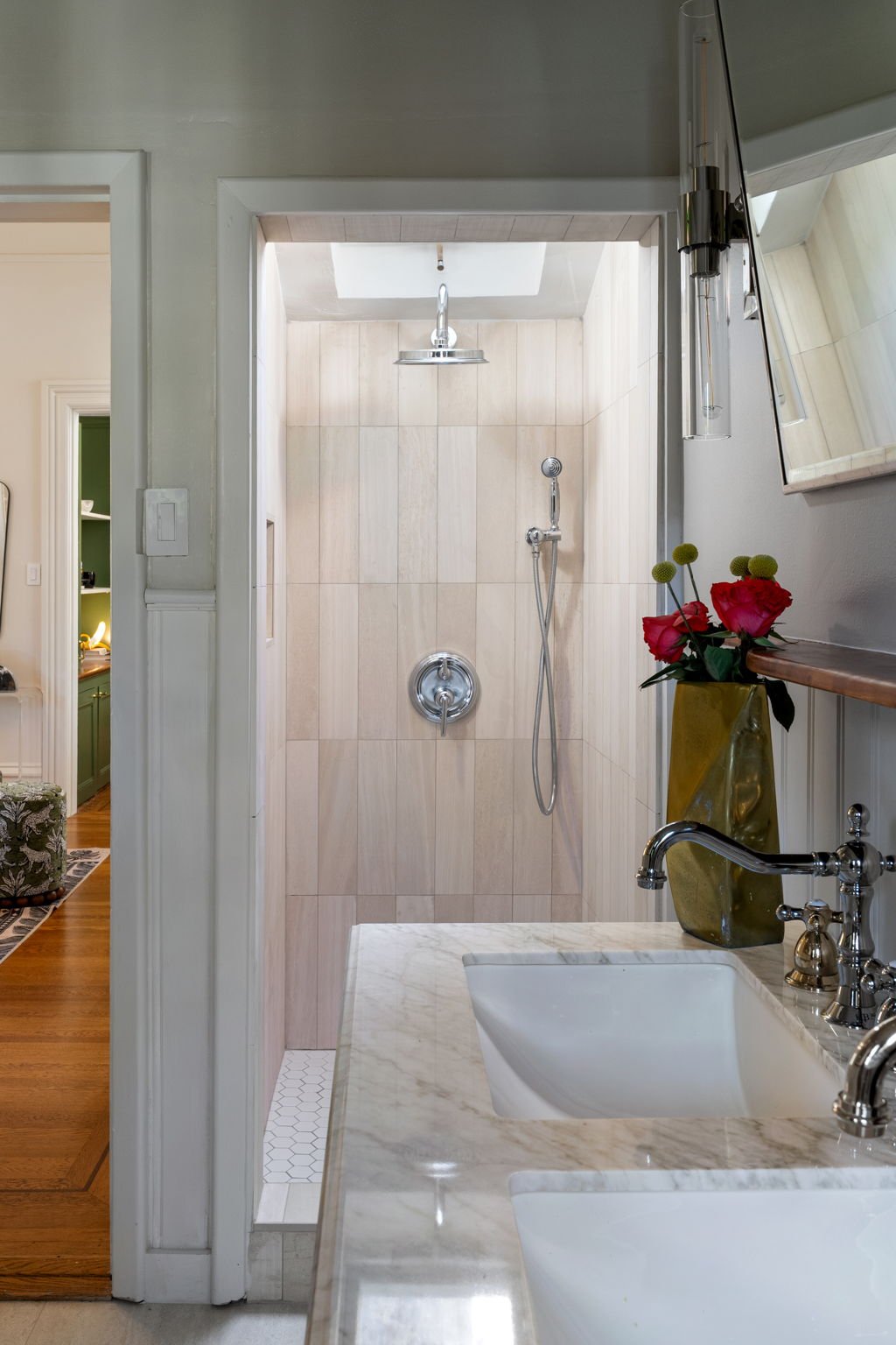 A bathroom with a walk-in shower featuring a ceiling shower head and a handheld shower head. There is a sunflower vase with red flowers on the side.