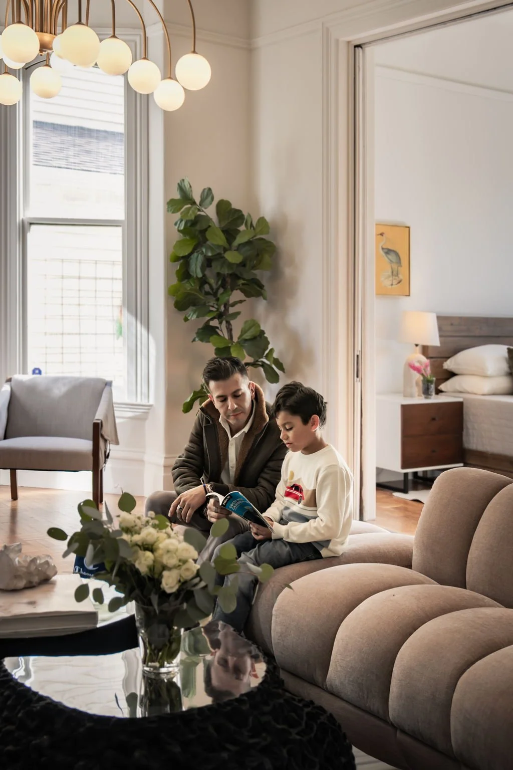 A man and a boy sitting on a beige sofa in a living room, looking at a magazine together. The room has a modern decor with a large window, a tall plant, and a bedroom visible in the background.