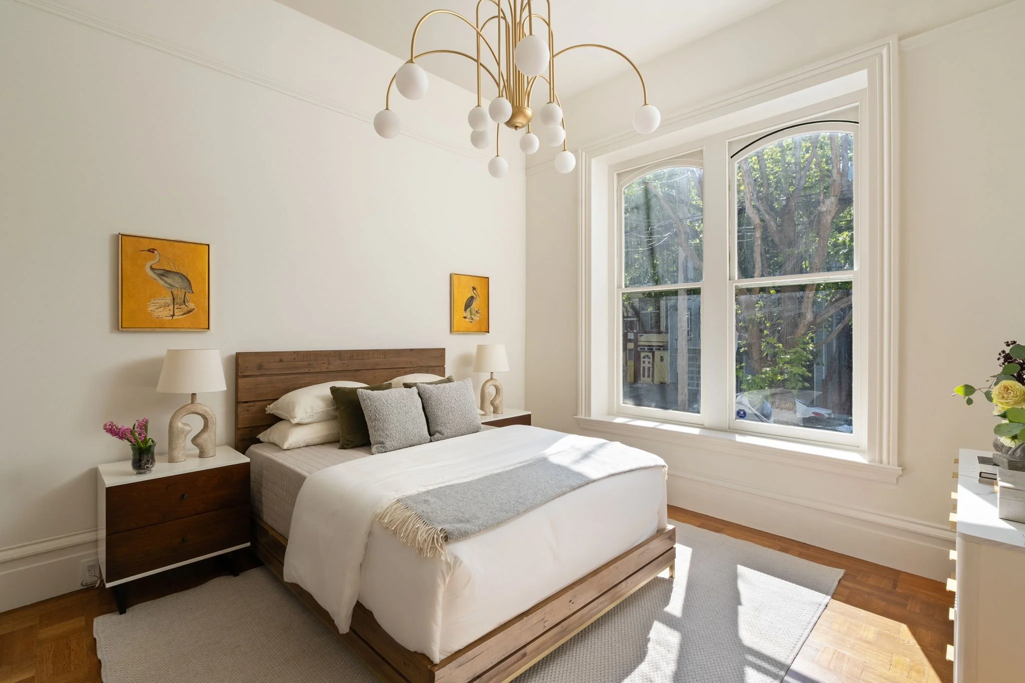 A bedroom with a large window, white walls, a wooden bed frame with white bedding, and two nightstands with lamps. Decorative animal artwork hangs above the bed, and a chandelier hangs from the ceiling.