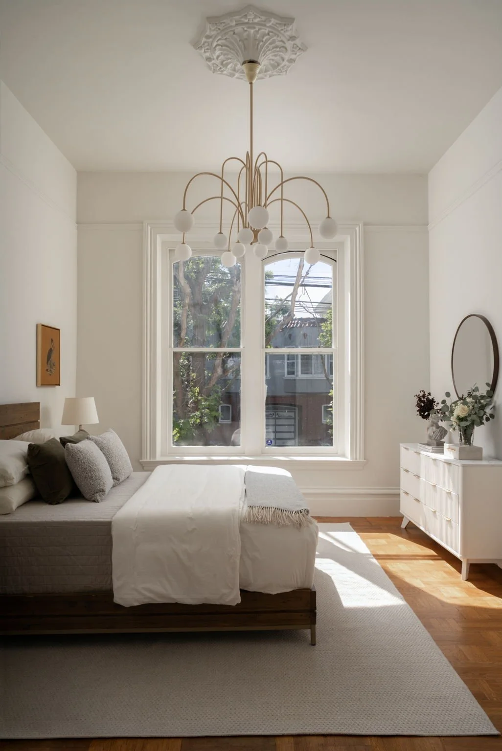 A bedroom with a wooden bed, white bedding, pillows, a nightstand with a lamp, a white dresser with a mirror and flowers, large window with trees outside, hardwood floor, and modern chandelier with globe lights.