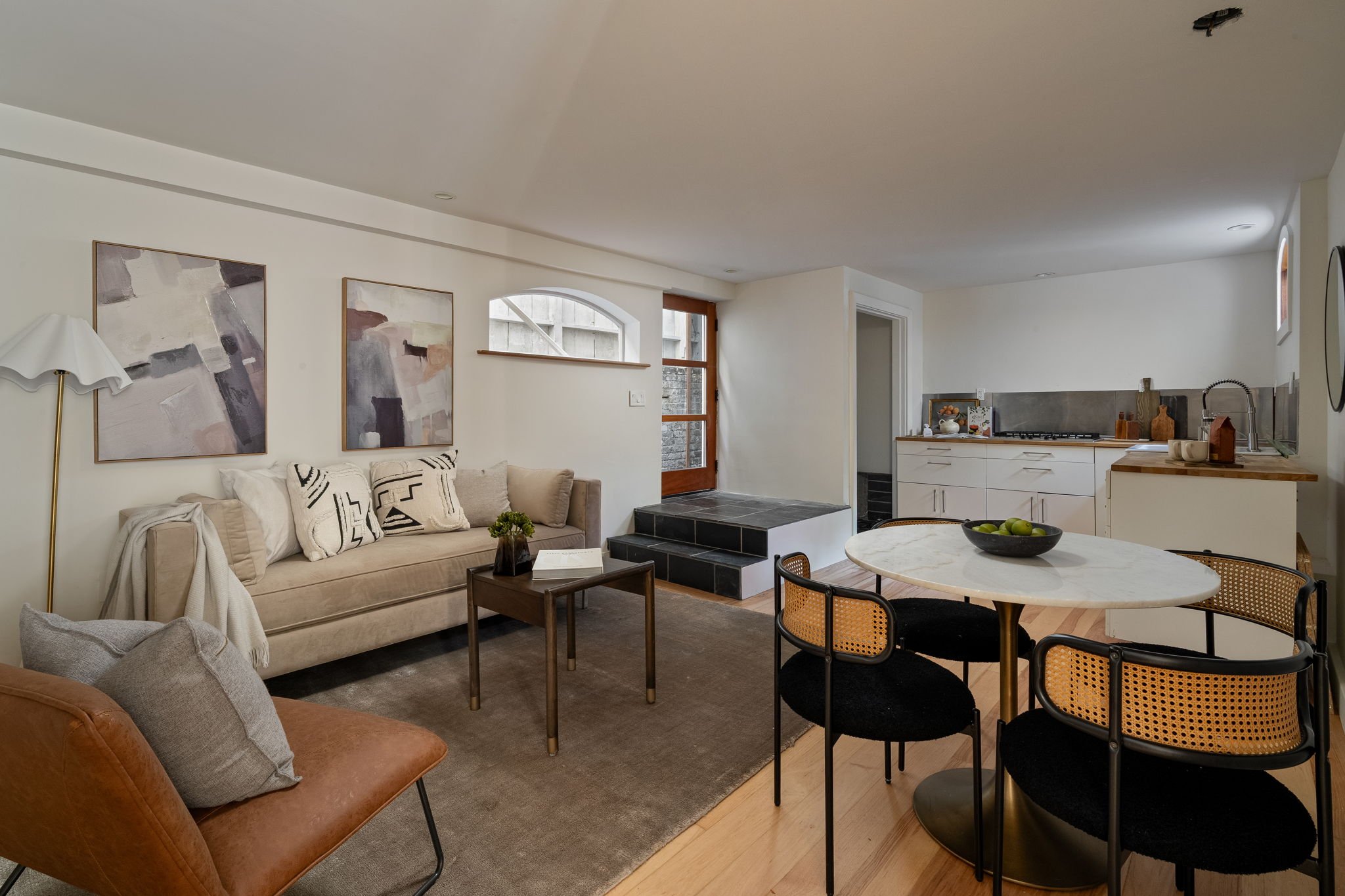 A living room and kitchen area with a beige sofa, abstract artwork on the wall, a small coffee table with a plant, and a round dining table with four rattan chairs. The kitchen has white cabinets, a backsplash, and a countertop with decorative items 