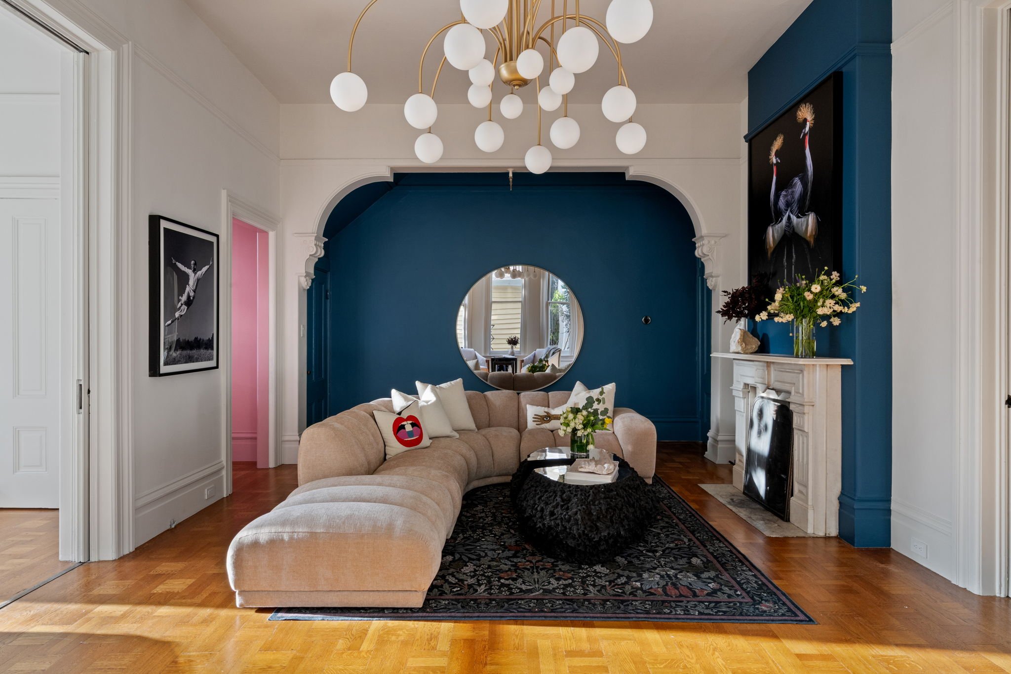 Living room with blue accent wall, beige curved sofa with decorative pillows, black patterned rug, white fireplace with floral arrangement and sculpture, round mirror on wall, hanging chandelier with white globes, framed black and white photo, hardwo