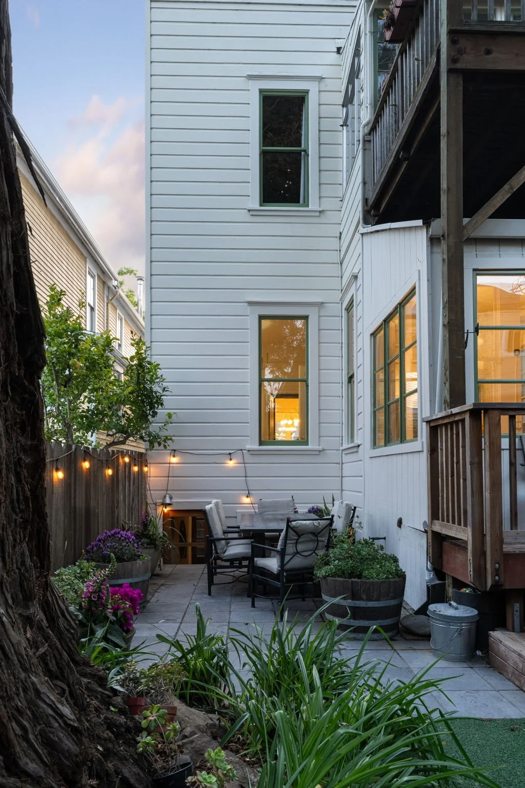 An outdoor patio area beside a white house with green window frames, decorated with string lights, potted flowers, and a wooden table with chairs, illuminated by evening light.