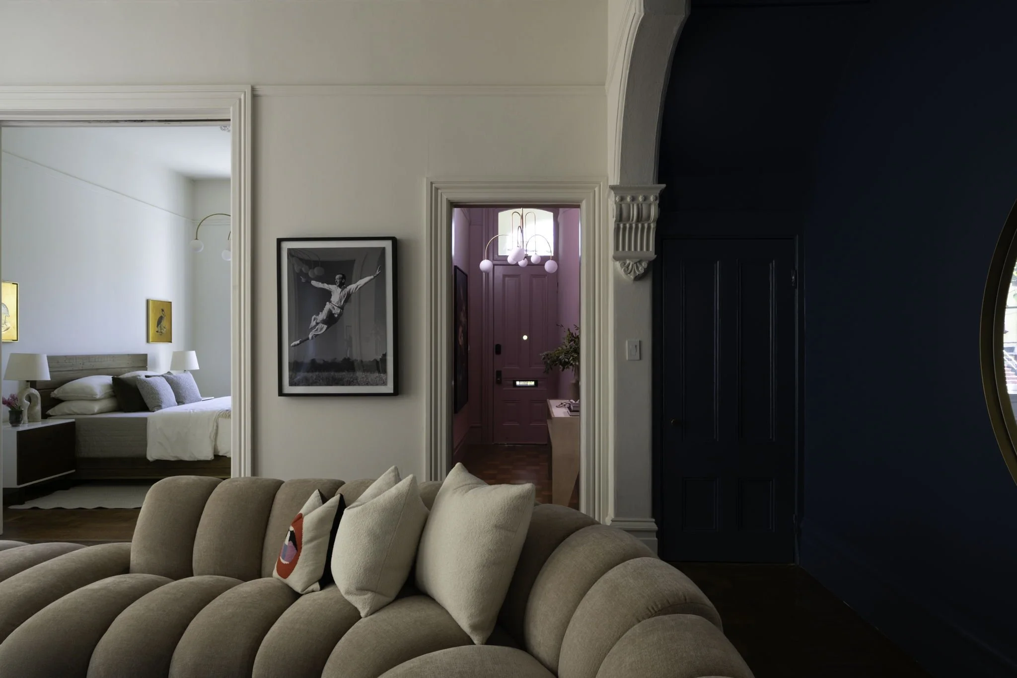 A living room with a beige tufted sofa, decorative pillows, a beige bedroom, framed artwork, and a pink hallway in the background.