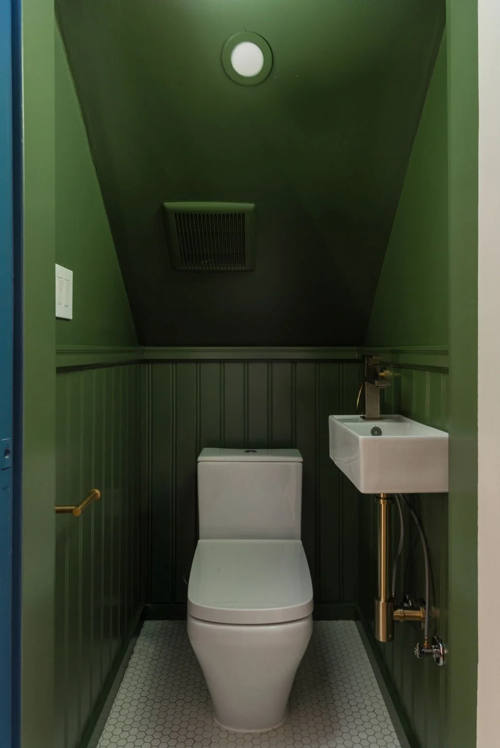 Small bathroom with dark green walls, a white toilet, a small wall-mounted sink with brass accents, a ceiling vent, and a white honeycomb tile floor.
