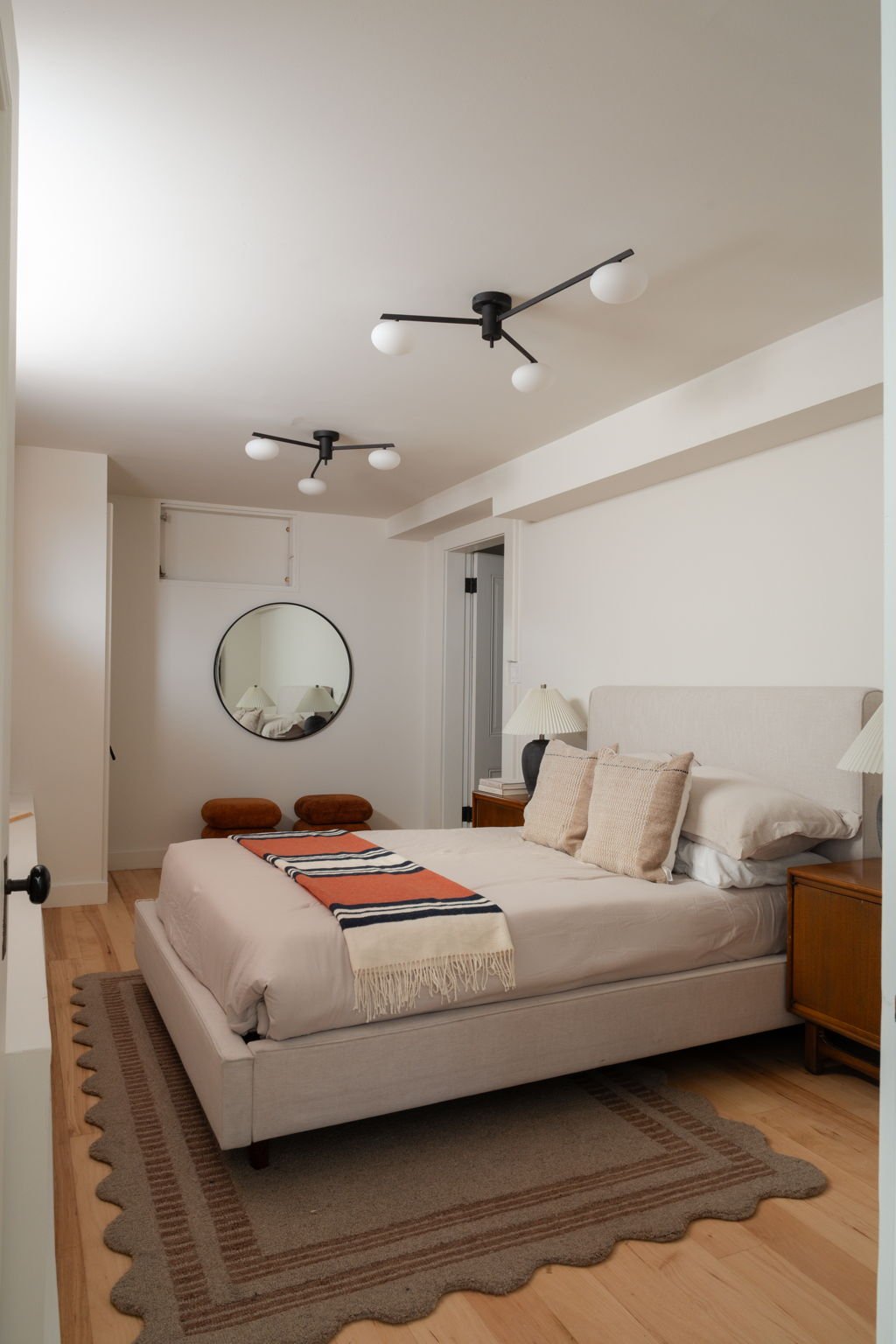 A modern bedroom featuring a white bed with multiple pillows, a striped blanket, wooden nightstands with lamps, a round mirror on the wall, and two brown ottomans under the mirror. The ceiling has two black modern light fixtures, and a gray area rug 