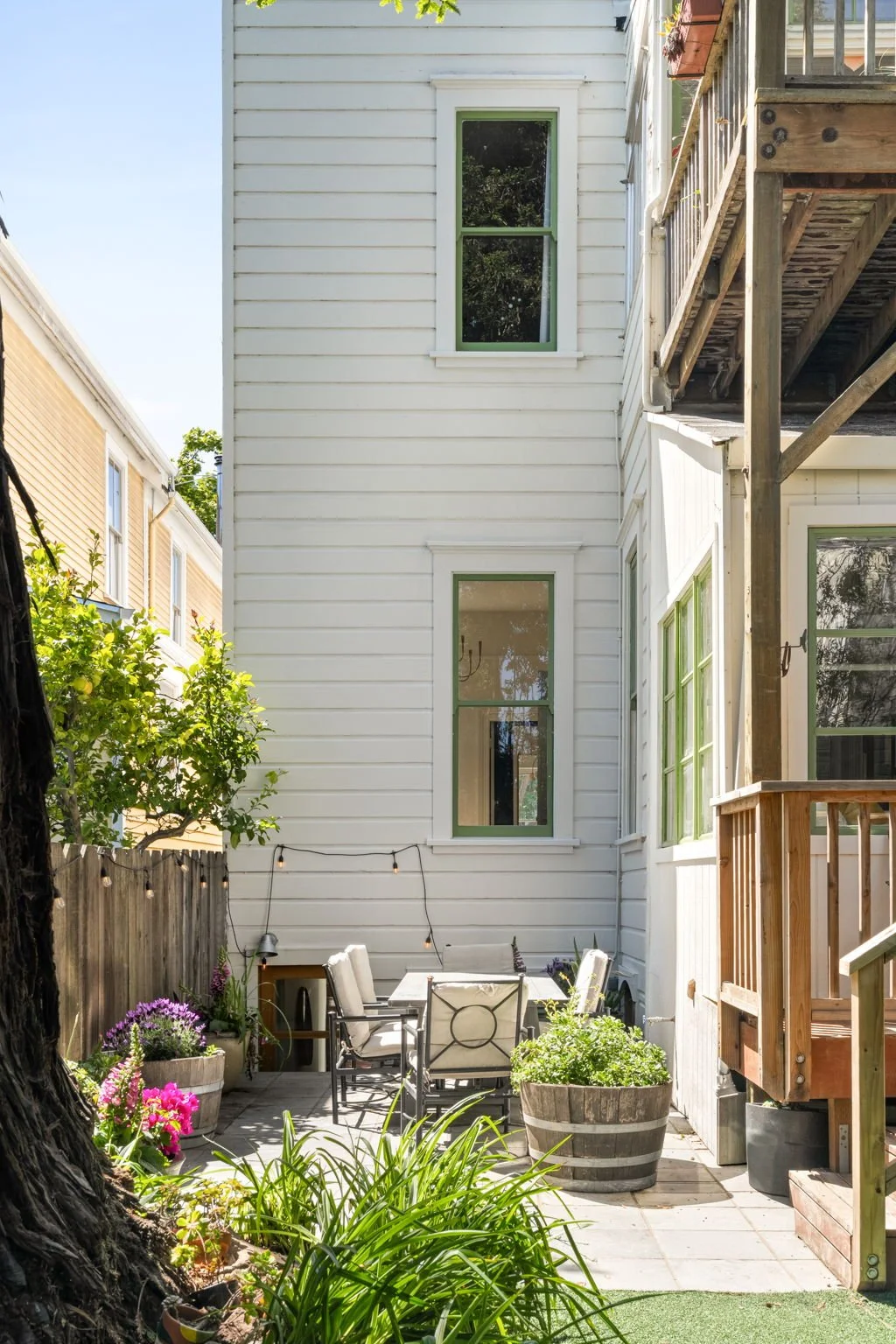 Small backyard patio with outdoor dining set, potted plants, string lights, surrounded by white and tan houses, green foliage.