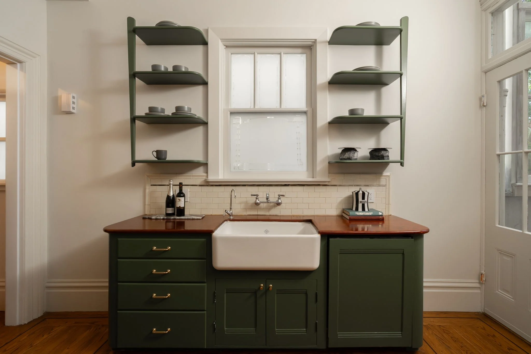 A vintage kitchen sink with a dark green cabinet base and a white porcelain basin, set against a cream wall with open green shelves above, featuring gray dishes and bowls. There are two liquor bottles on the counter, and a window in the background