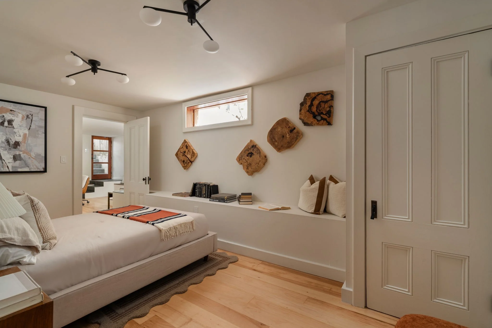 A cozy bedroom with white walls, a low window, and wooden decor pieces on the wall above the bed.