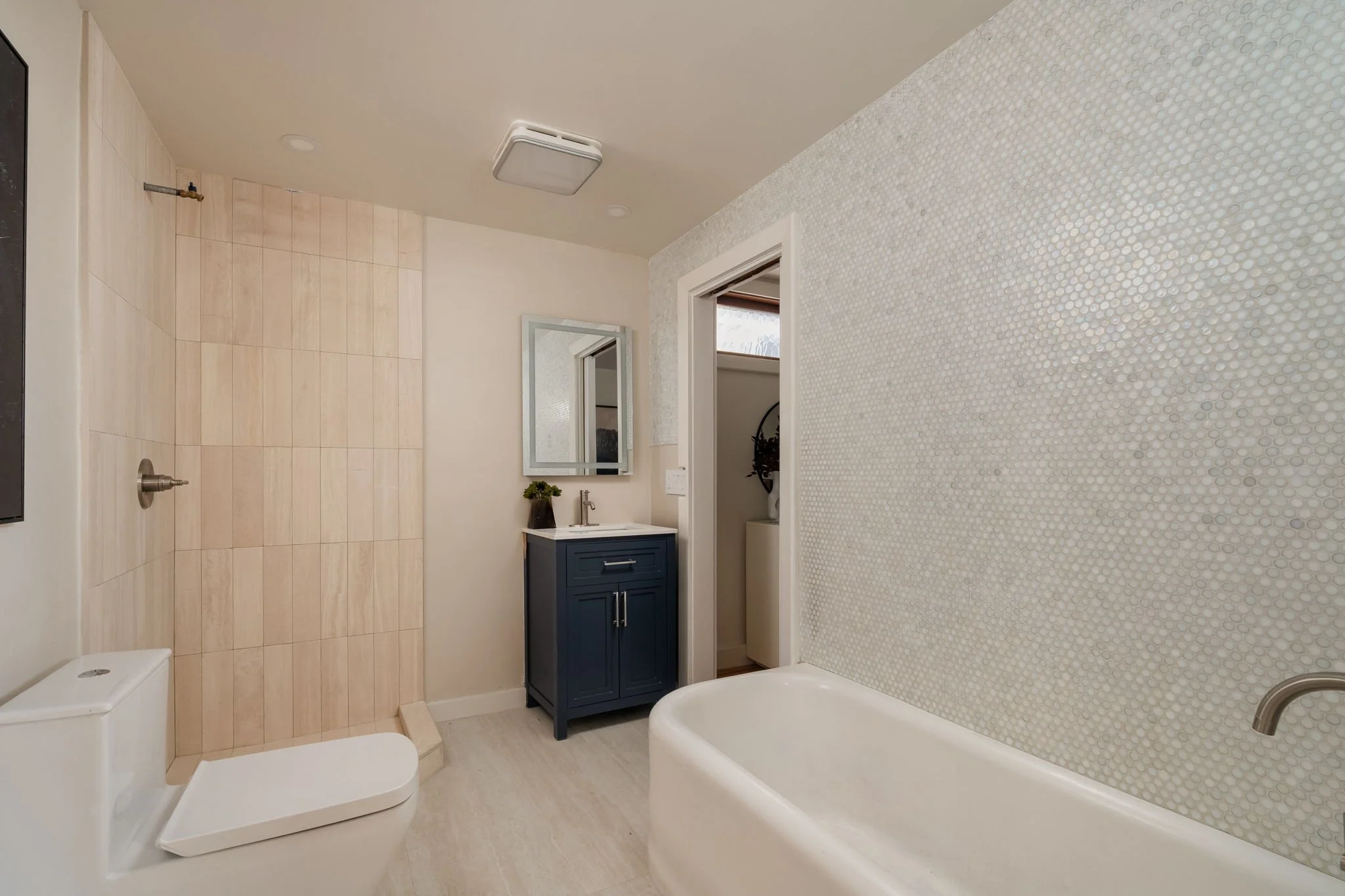 A bathroom with a beige tile floor and a textured mosaic wall, featuring a bathtub, a blue vanity with a mirror, a small plant, a ceiling light, and a doorway leading to a toilet area.