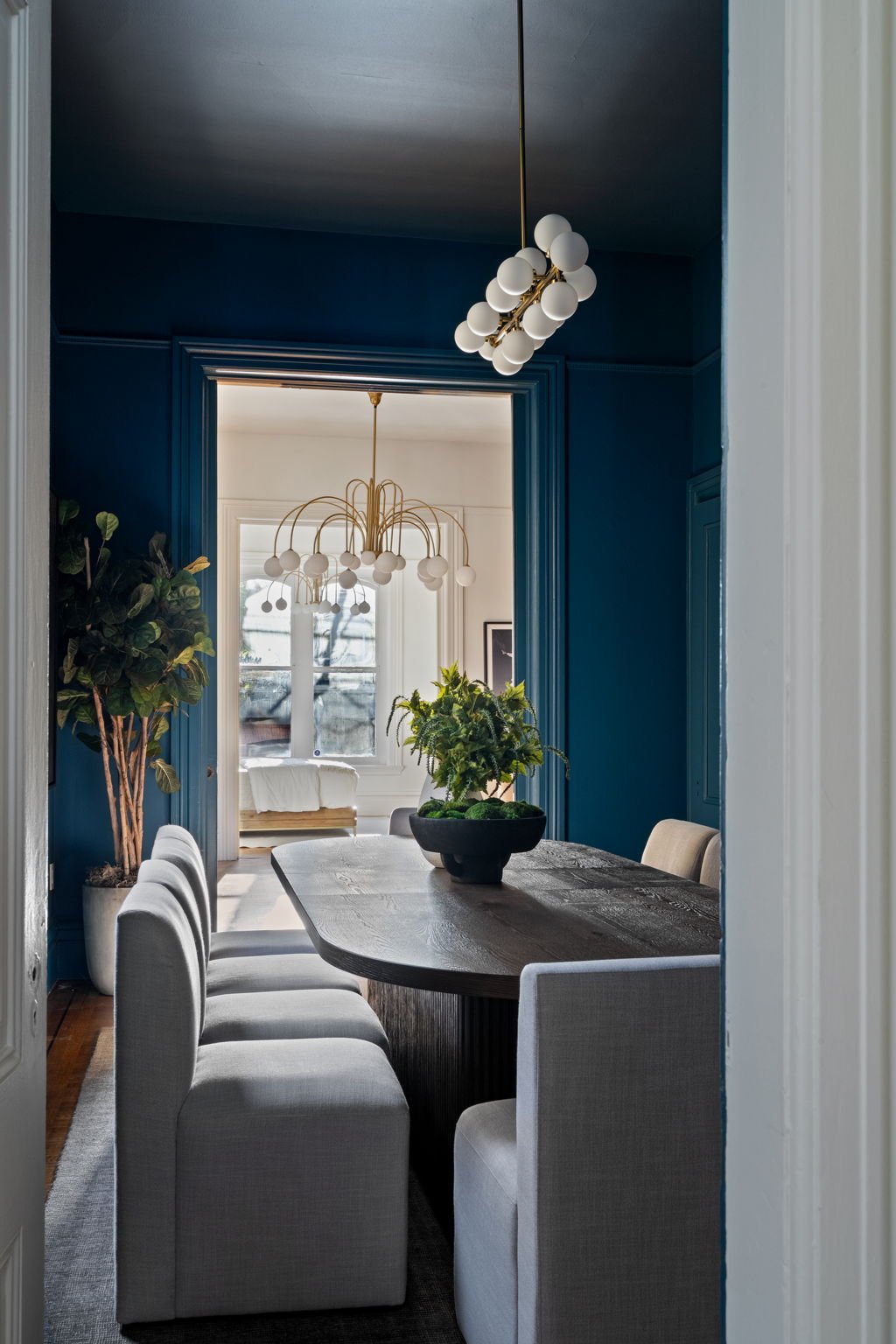 View of a dining room seen through a doorframe, with white upholstered chairs around a dark wooden table, green plants, and two modern chandeliers, one in the foreground with white globes and gold accents, and another in the background with white glo