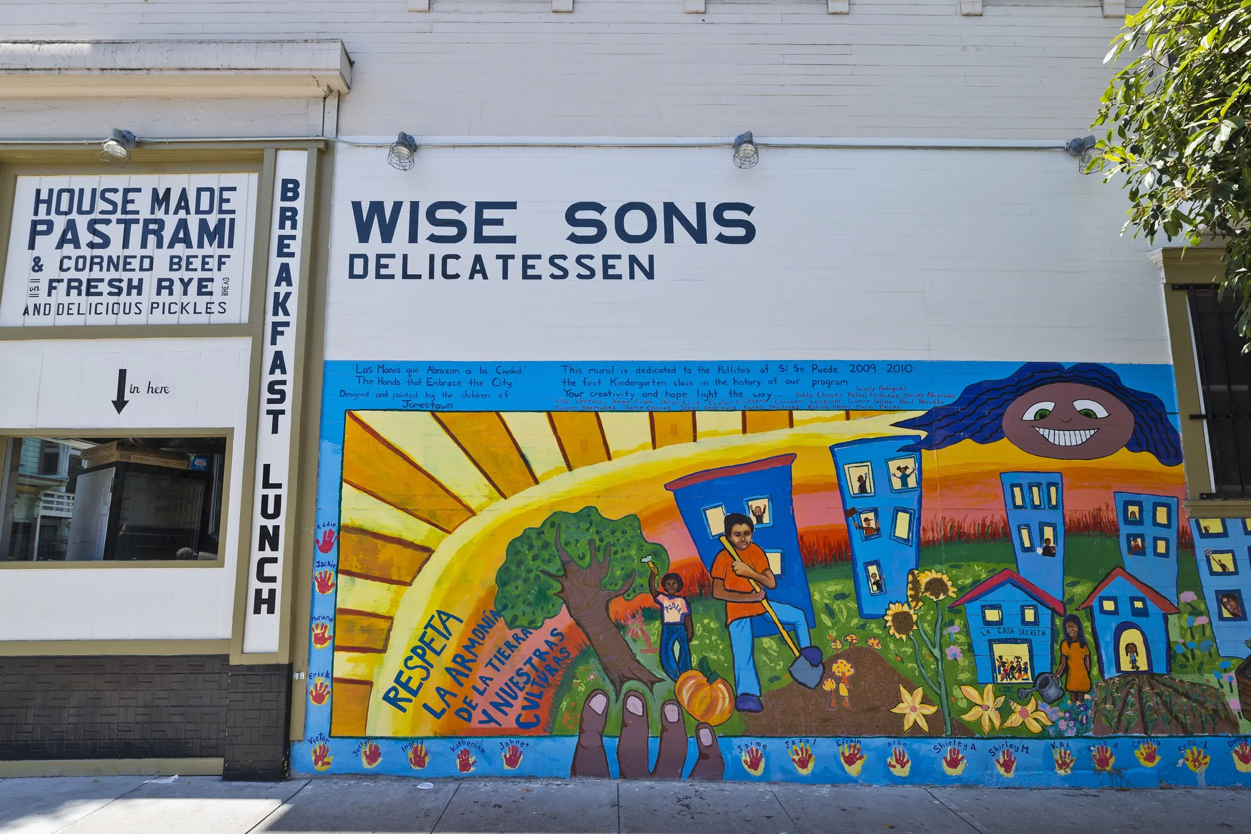 Colorful mural depicting children playing in a neighborhood with trees and houses, with a bright sun in the background. The mural includes handwritten text about community and education.