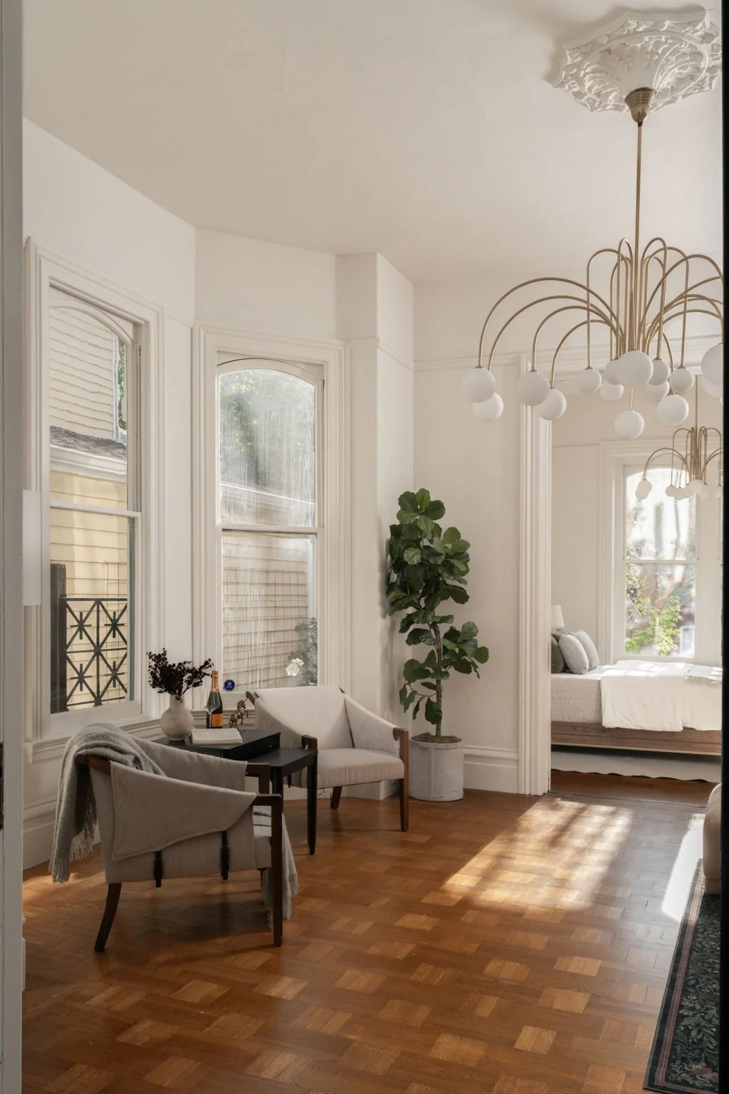 A bright living room with large arched windows, a potted fiddle leaf fig plant, two beige armchairs, a small black side table with a vase and a bottle, and a view into a sunlit bedroom with a daybed.