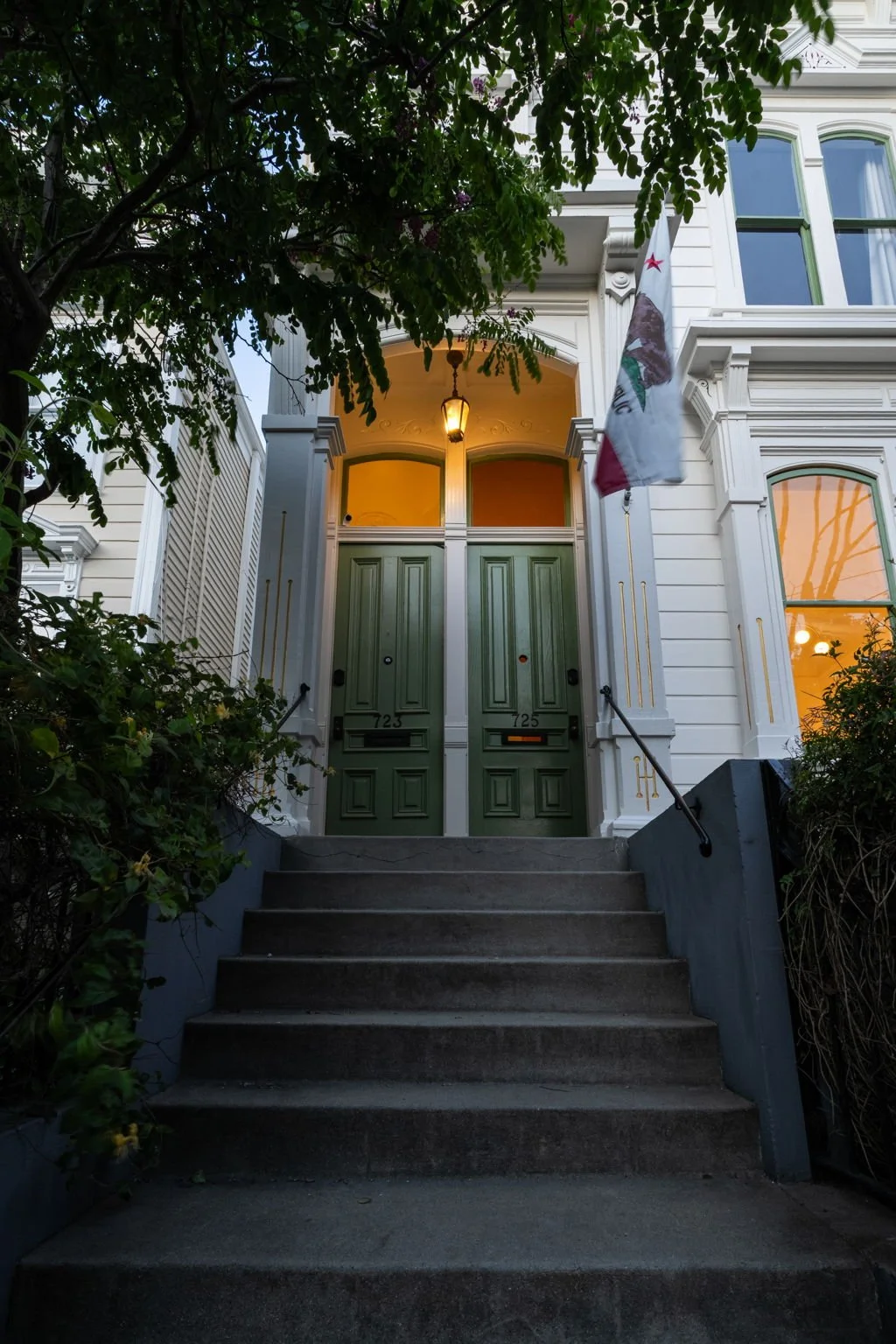 Front entrance of a Victorian-style house with two green doors, stairway, overhanging porch light, flag, and surrounding greenery, in evening light.