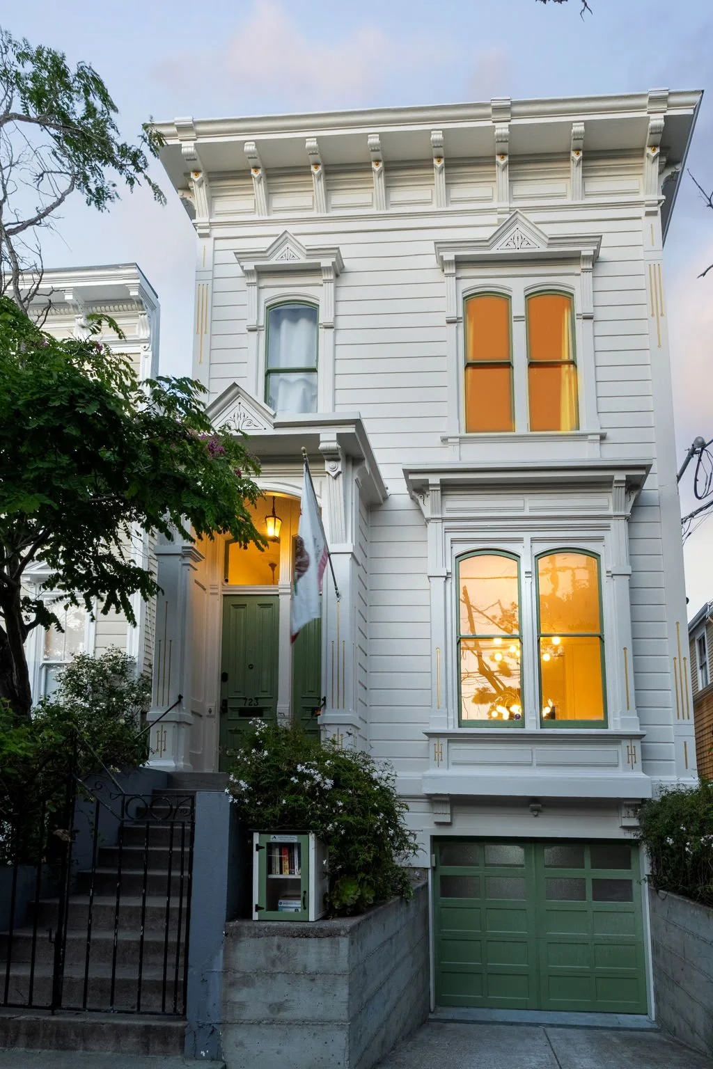 A white Victorian-style house with green door and window frames, multiple windows with orange and white curtains, a flag hanging by the entrance, a small garden with plants, and a closed green garage door, during dusk.