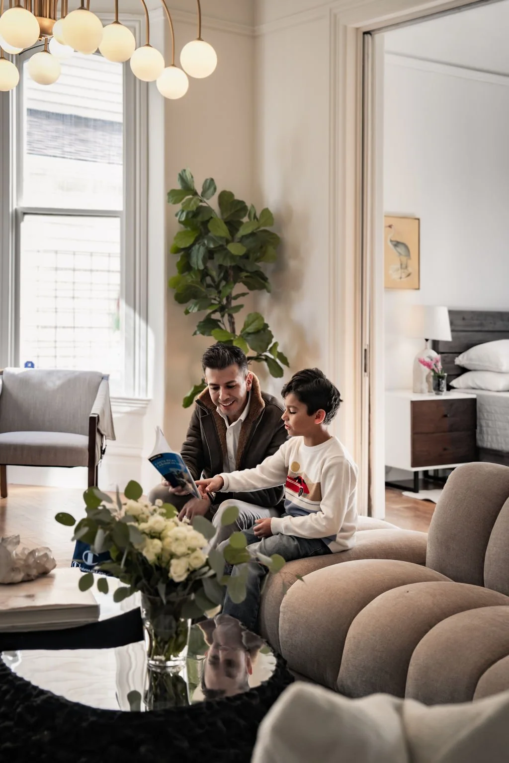 A man and a boy sitting on a beige sofa in a living room, looking at a book or magazine together, with a flower arrangement on a black coffee table in the foreground.