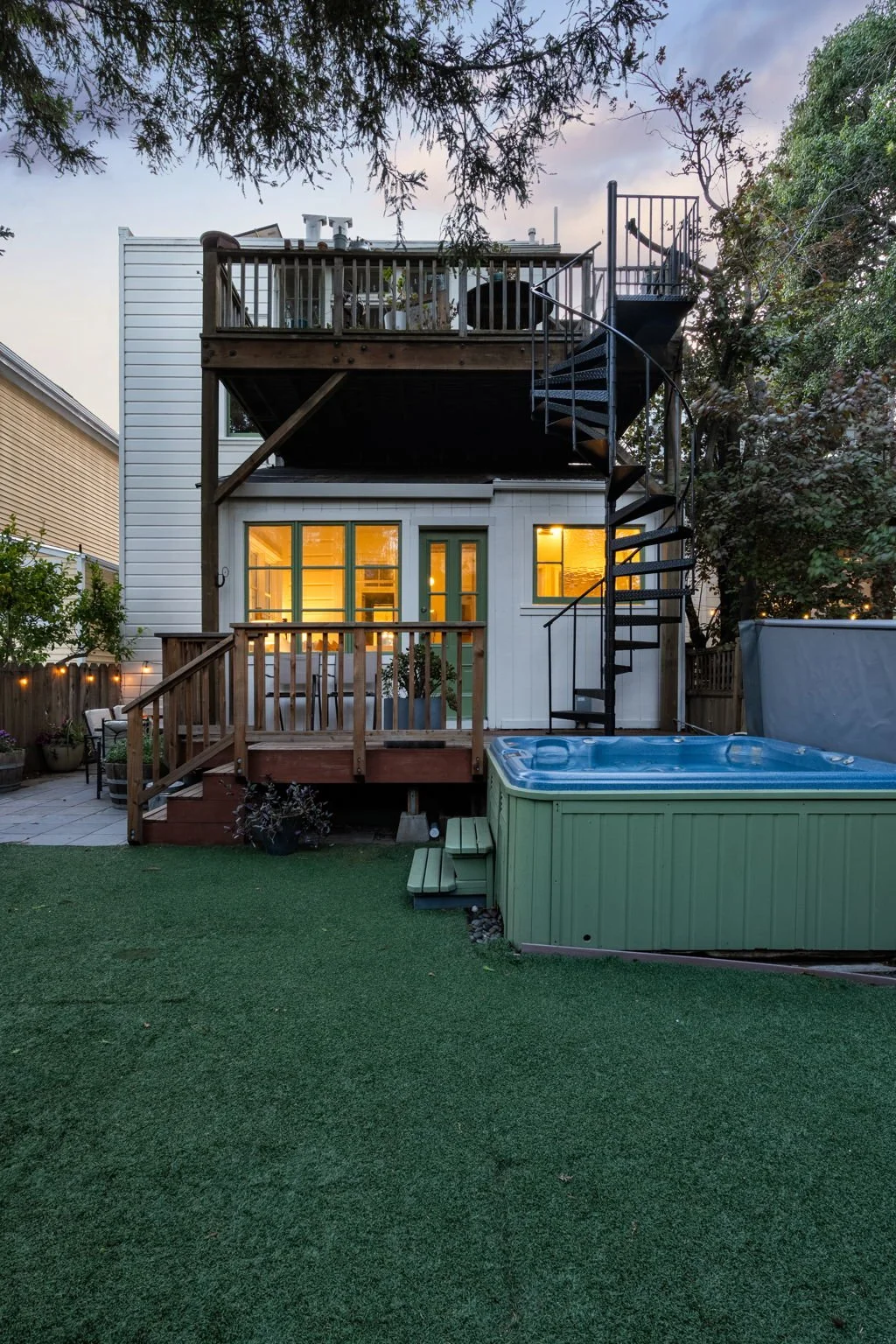 Backyard view of a house with two levels, illuminated windows, a spiral staircase on the right, a wooden deck on the second floor, and a hot tub in the foreground on the right.