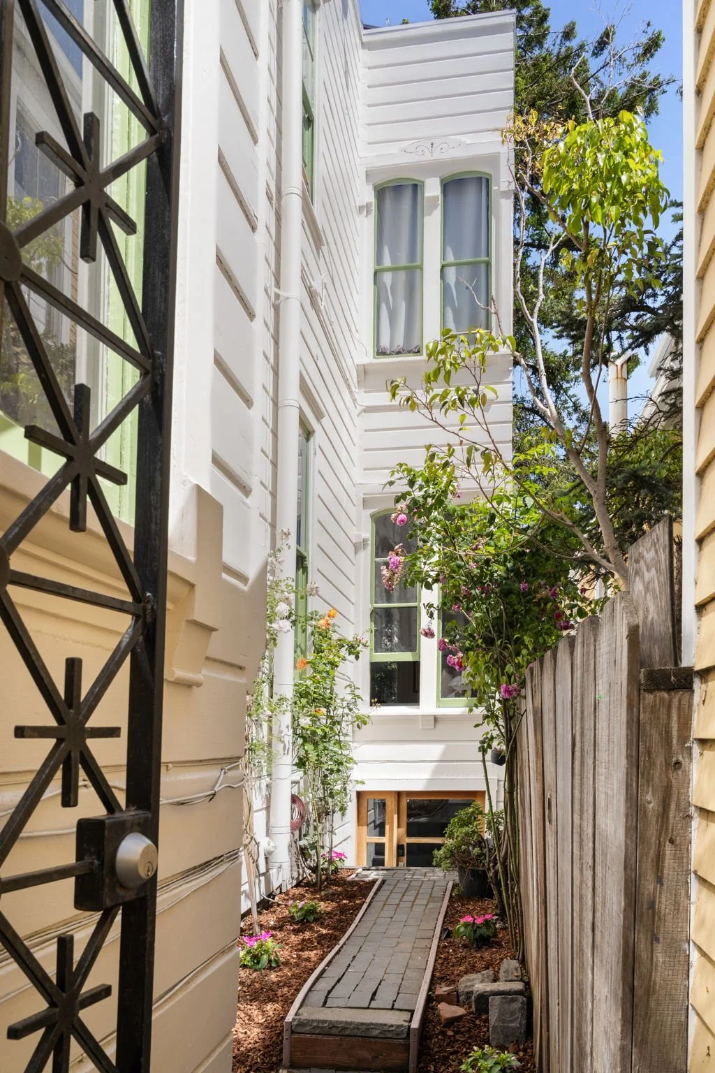 A narrow garden alley between two houses with wooden fencing and lush green trees, a small stone pathway, and flowering plants.