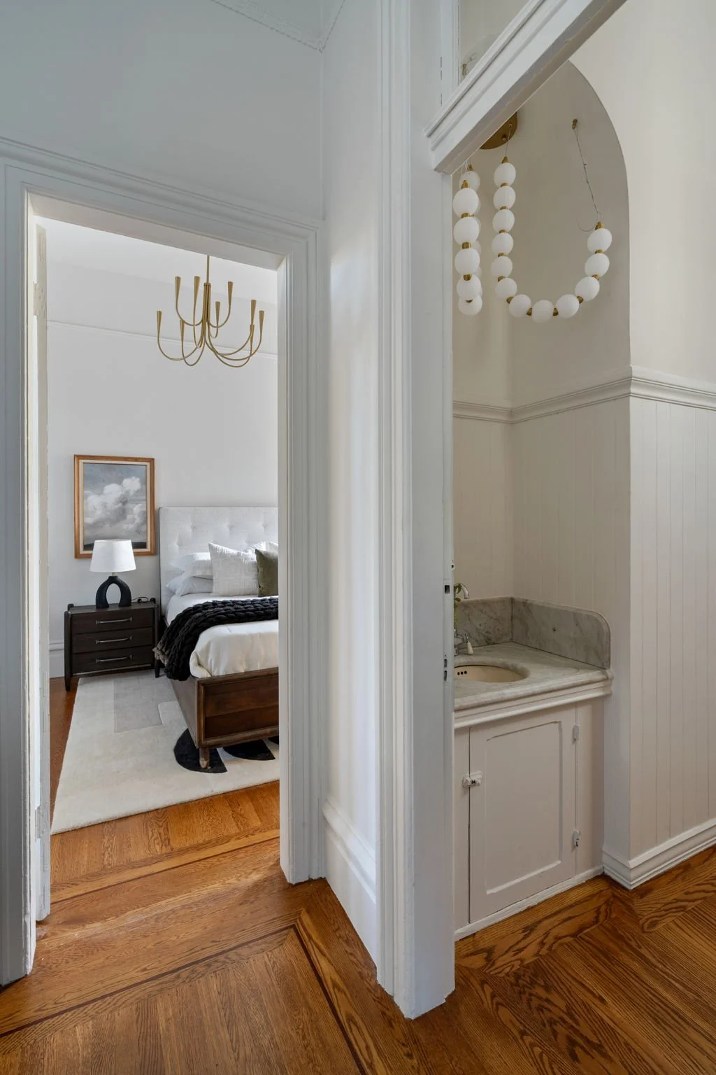 A view from a hallway into a bedroom with a white bed, a black nightstand, a white and black rug, and artwork on the wall. The hallway has a small vintage sink with a marble countertop and a decorative hanging light made of white spherical beads.
