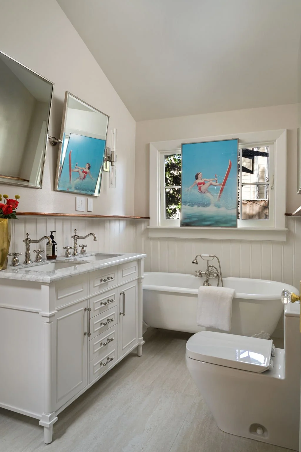 A bright bathroom with a white vanity, a bathtub, a toilet, and a window with seashell artwork mirroring beach scenes.