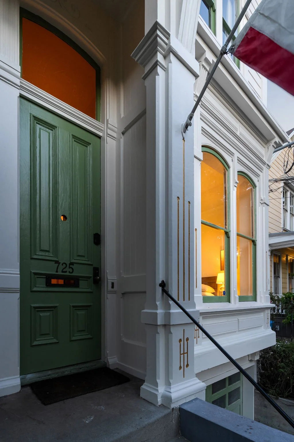 Close-up of a Victorian-style house with a green door marked 725, large windows with orange curtains, and a lit interior visible through the window. A black handrail runs along a step near the door, and part of a red, white, and gray awning is visibl