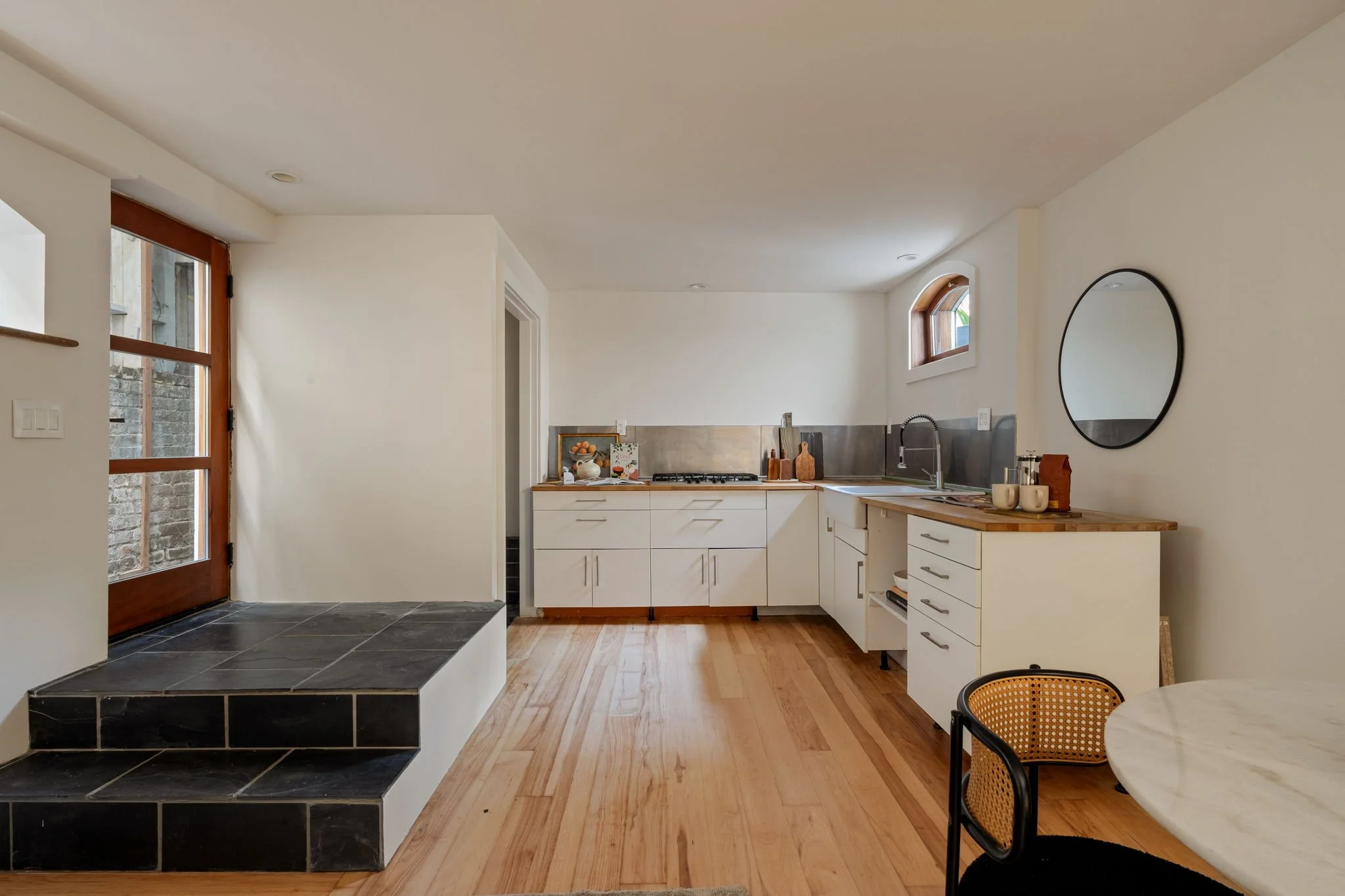 A modern kitchen with white cabinetry, a backsplash, and wooden accents, featuring small above-window openings in the wall, and a round mirror on the wall. There is a small raised platform with black tiles at the entrance.