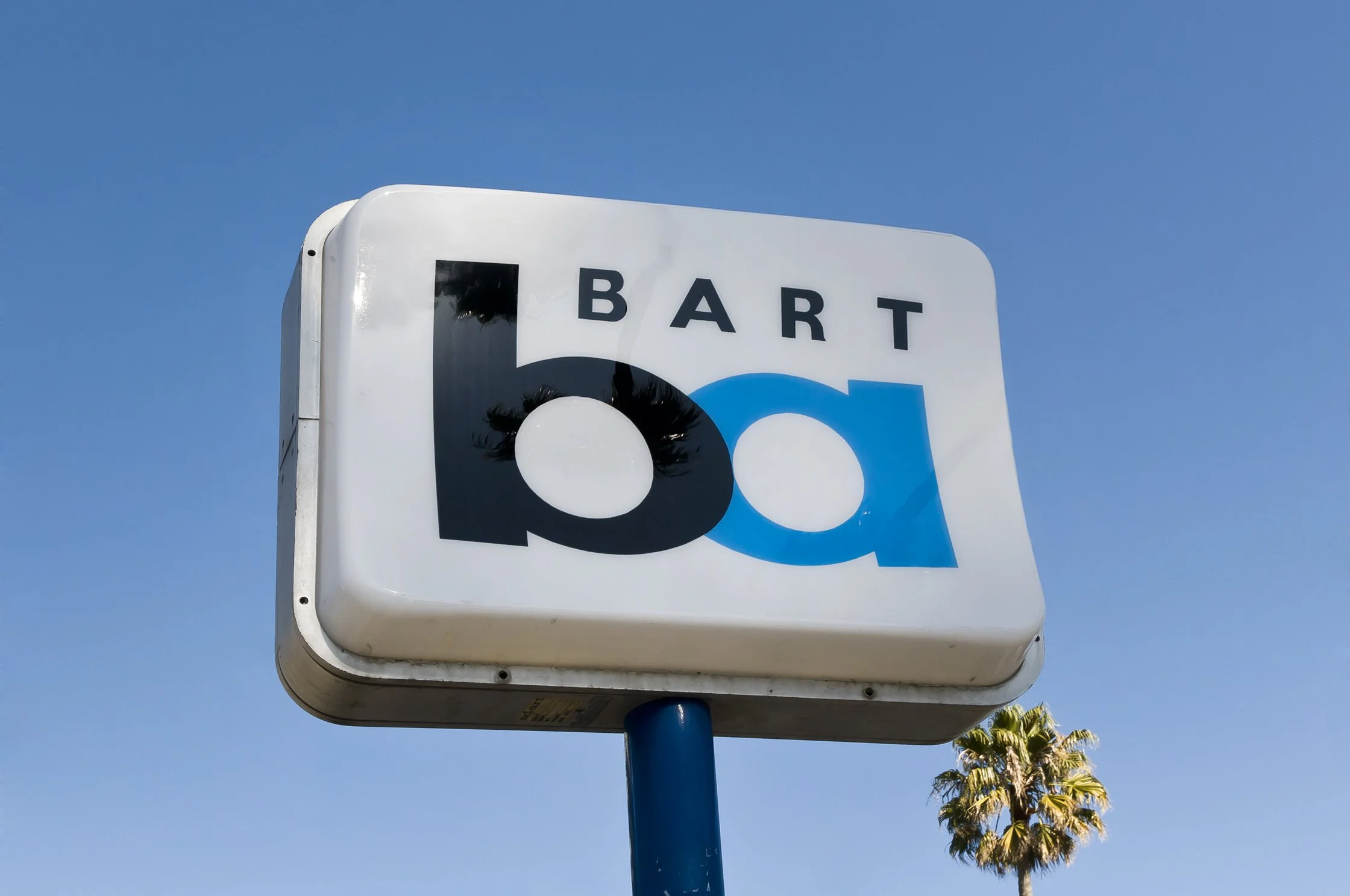 A road sign with the word "BART" and a circular graphic, mounted on a blue pole, against a clear blue sky with palm trees in the background.