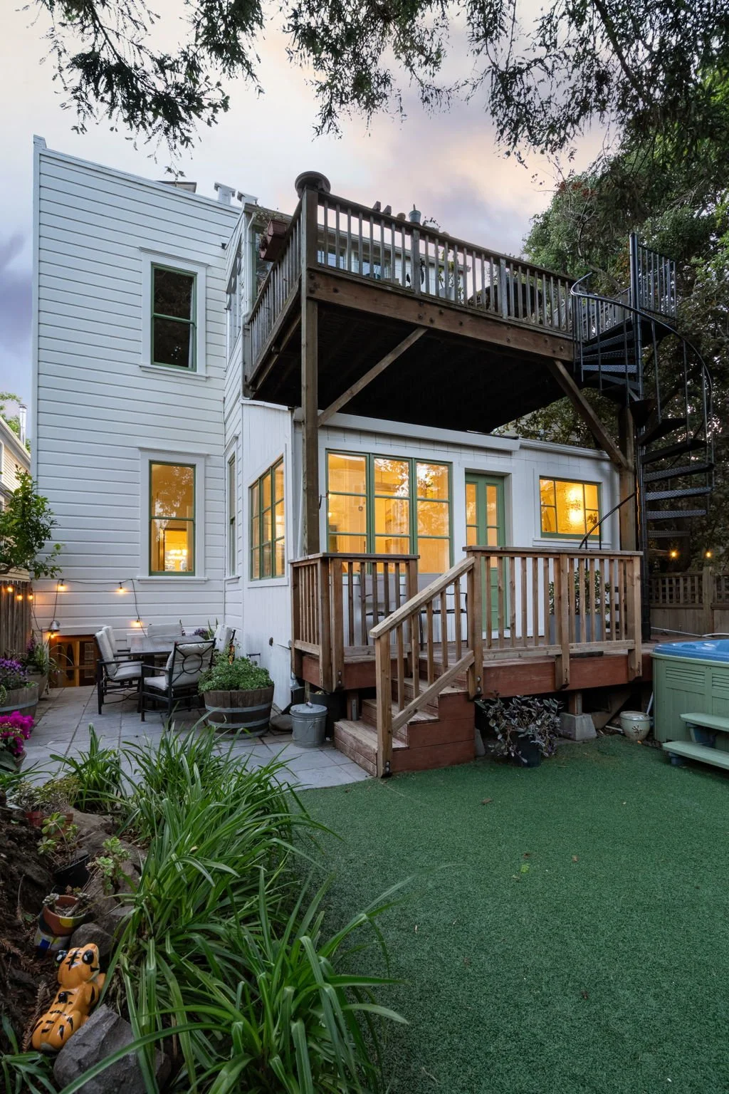 The back of a two-story white house with a wooden deck on the upper level and a small porch on the lower level, surrounded by a garden with plants, outdoor furniture, and a small artificial grass yard, during evening with warm indoor lights.