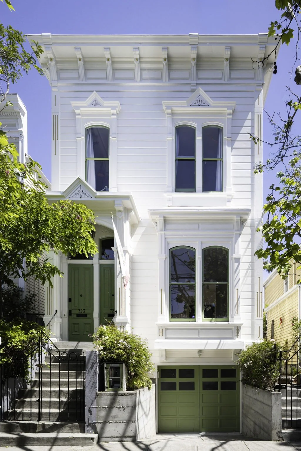 A white three-story Victorian-style house with green doors and window frames, surrounded by trees and shrubs, under a clear blue sky.