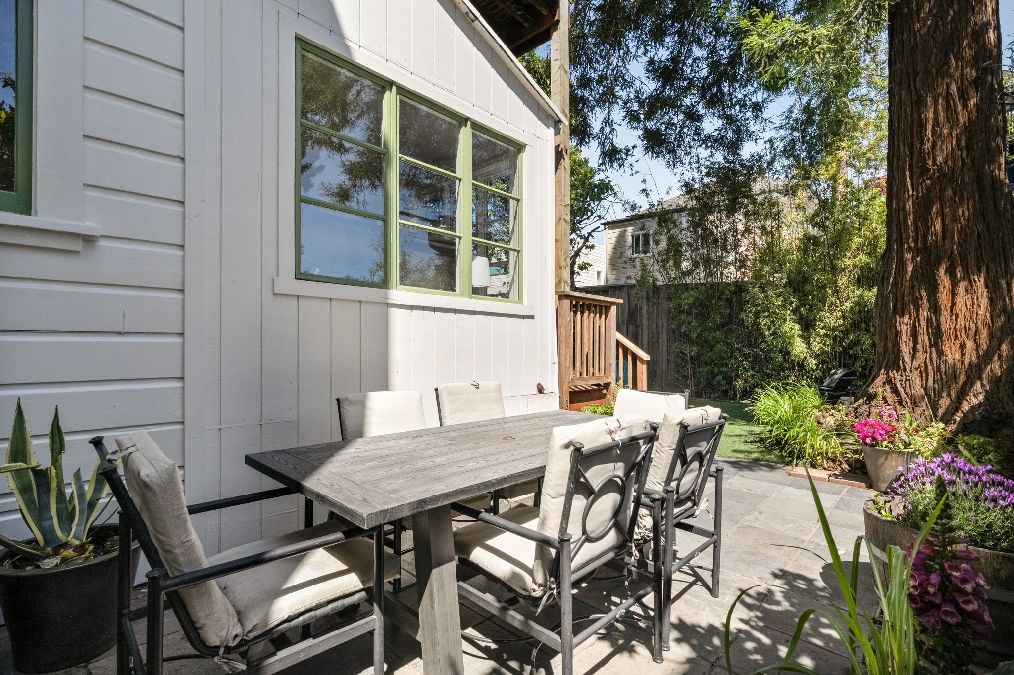 A backyard patio with a wooden dining table and metal chairs with cushions, surrounded by green plants and flowers, with a large tree and neighboring houses visible in the background.