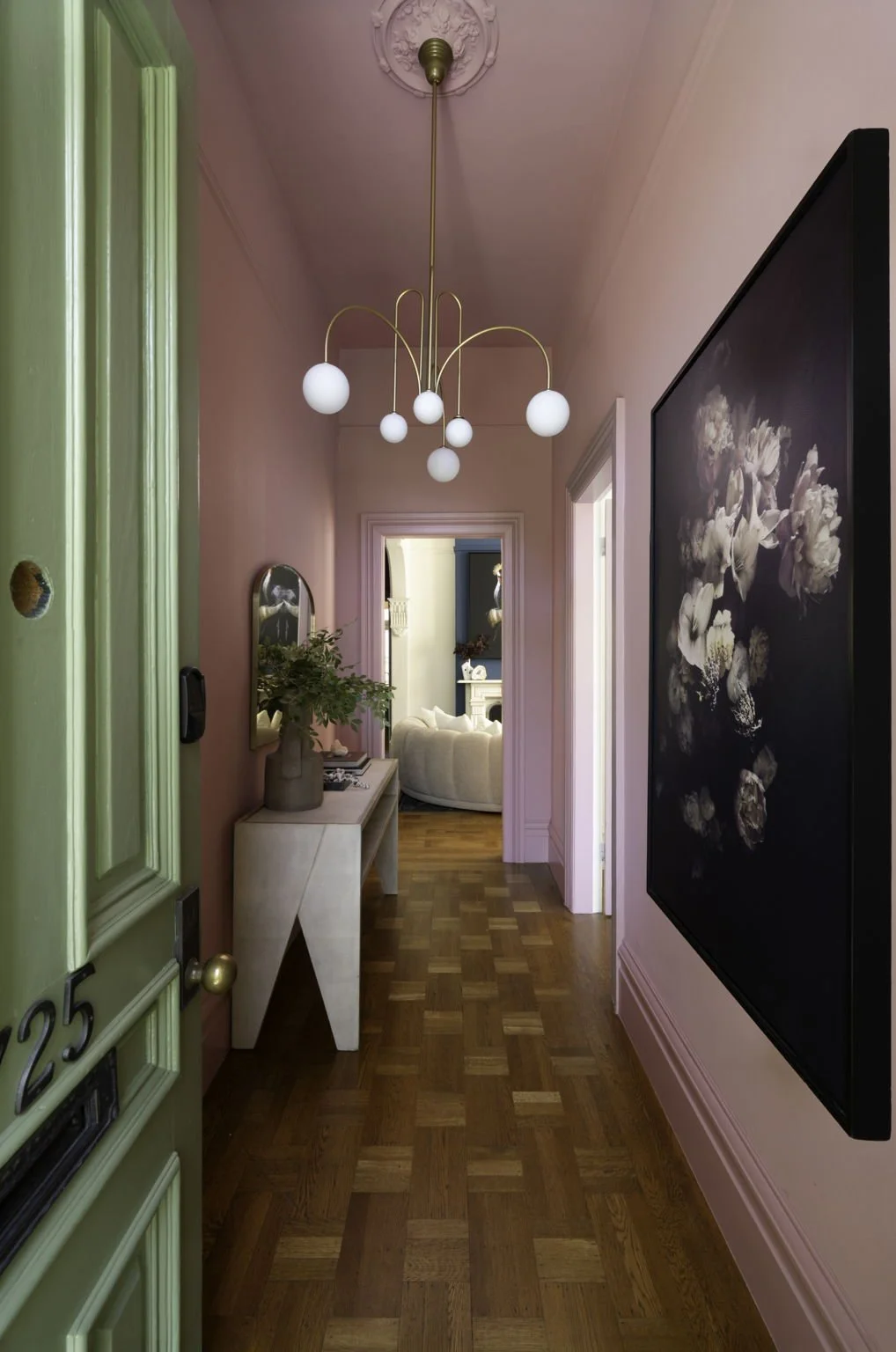 Interior view of a hallway with pink walls, hardwood flooring, a modern chandelier with white globes, a white console table with a vase and greenery, and a large floral painting on the wall.