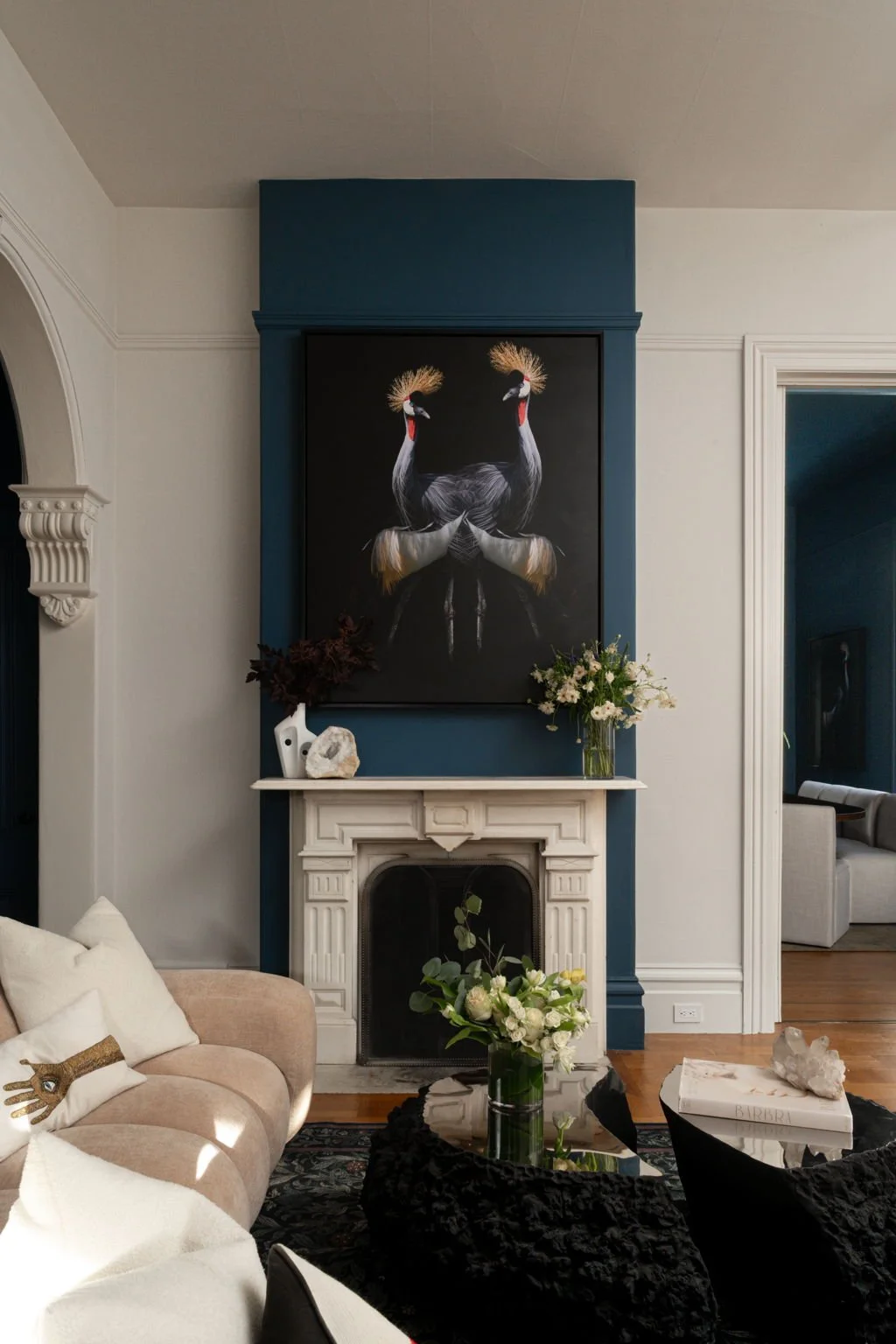 Living room with a white fireplace, large black and white painting of two cranes, and a beige sofa with pillows. A glass vase with white flowers sits on a wooden table in front of the fireplace, and a smaller black table holds a large book and a deco