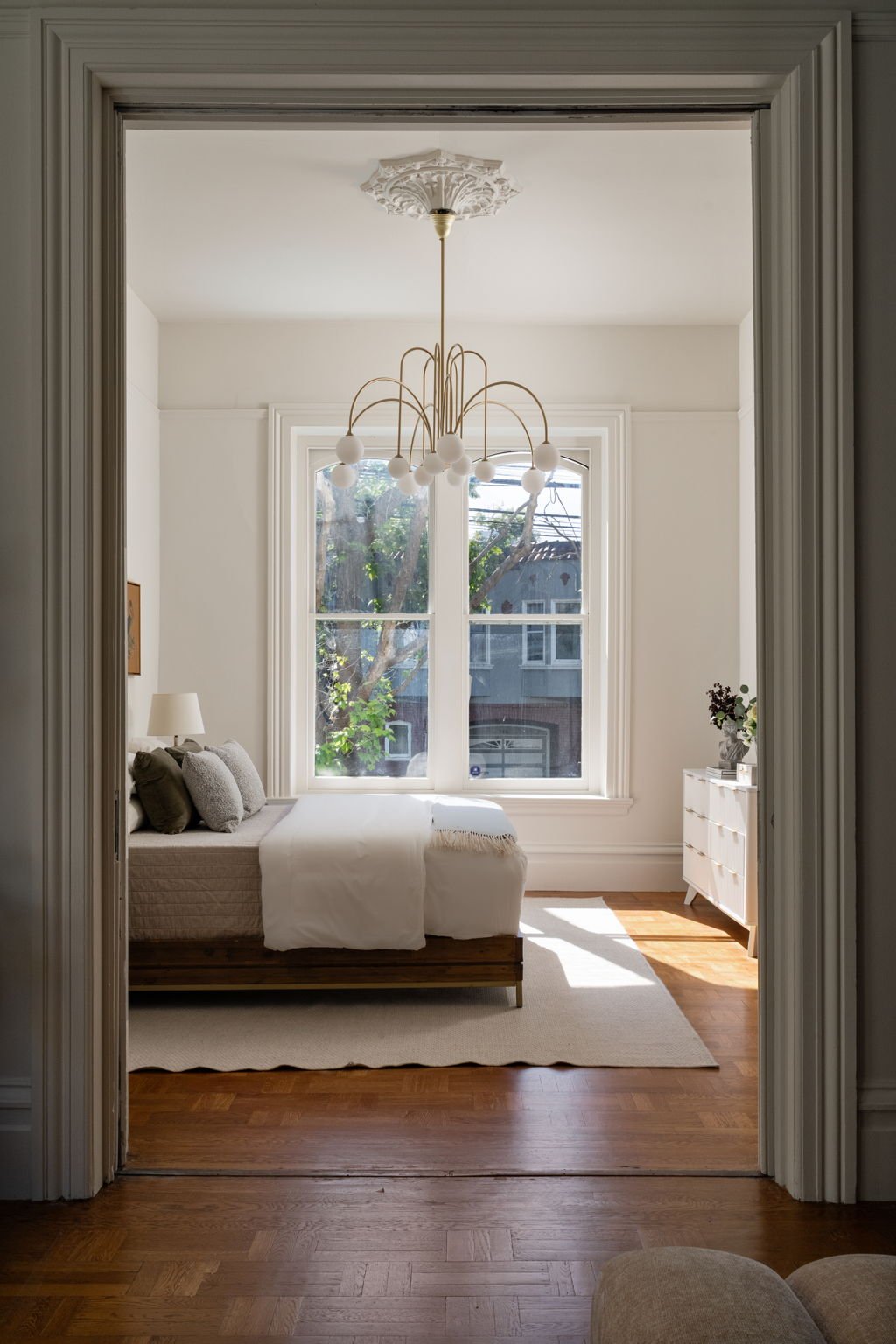 Bright bedroom with large window overlooking tree, white walls, bed with gray pillows, wooden bed frame, white bedding, white dresser with flowers, and modern chandelier.