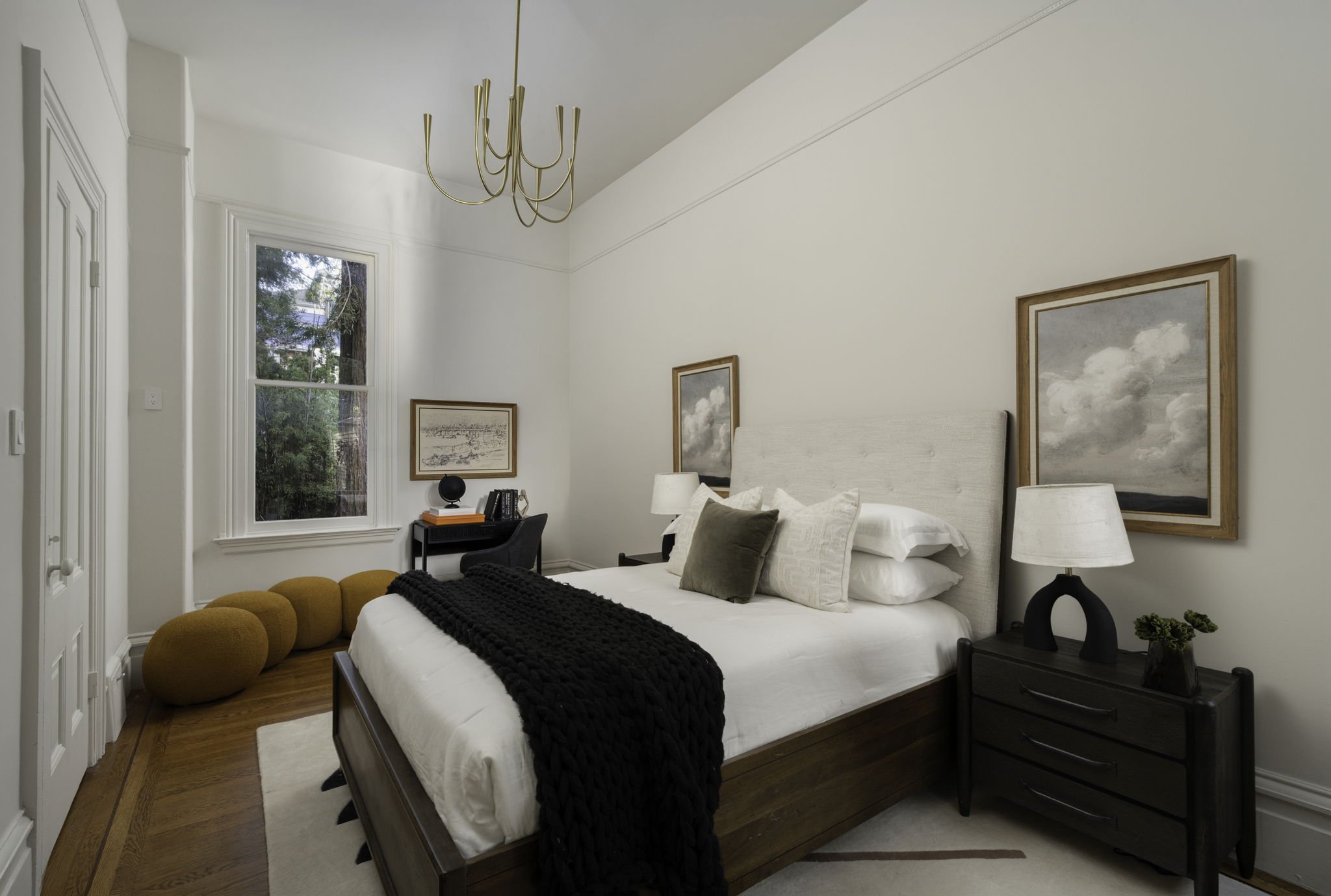 A bedroom with a white bed, beige headboard, black throw blanket, and several pillows. There are two nightstands with lamps, framed artwork on the wall, a window with a view of trees, a ceiling chandelier, and a mustard-colored ottoman with multiple 