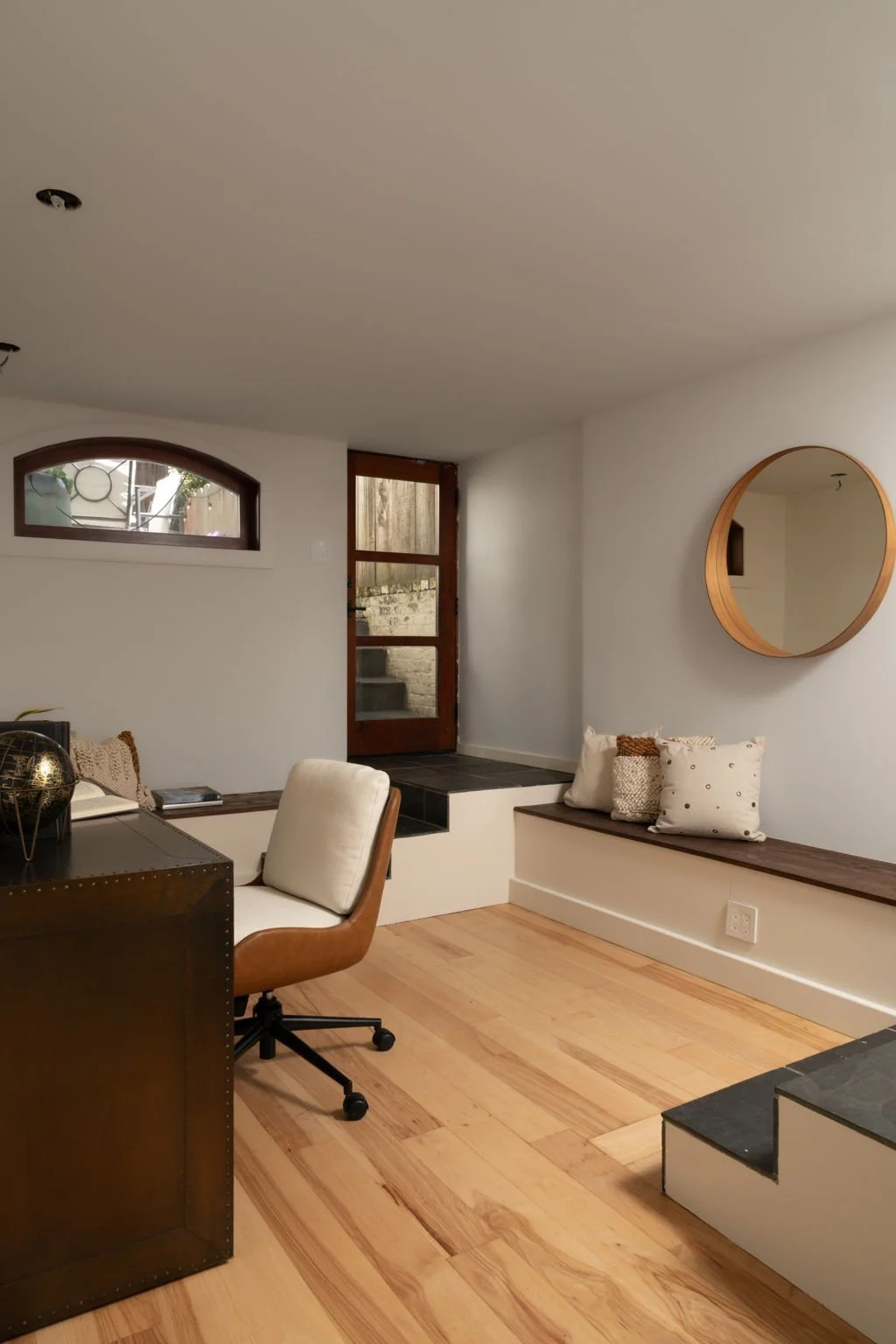 A cozy living room with light-colored wooden flooring, a small window, stairs leading outside, a wooden bench with decorative pillows, a round wall mirror, and a desk chair with a globe and papers.