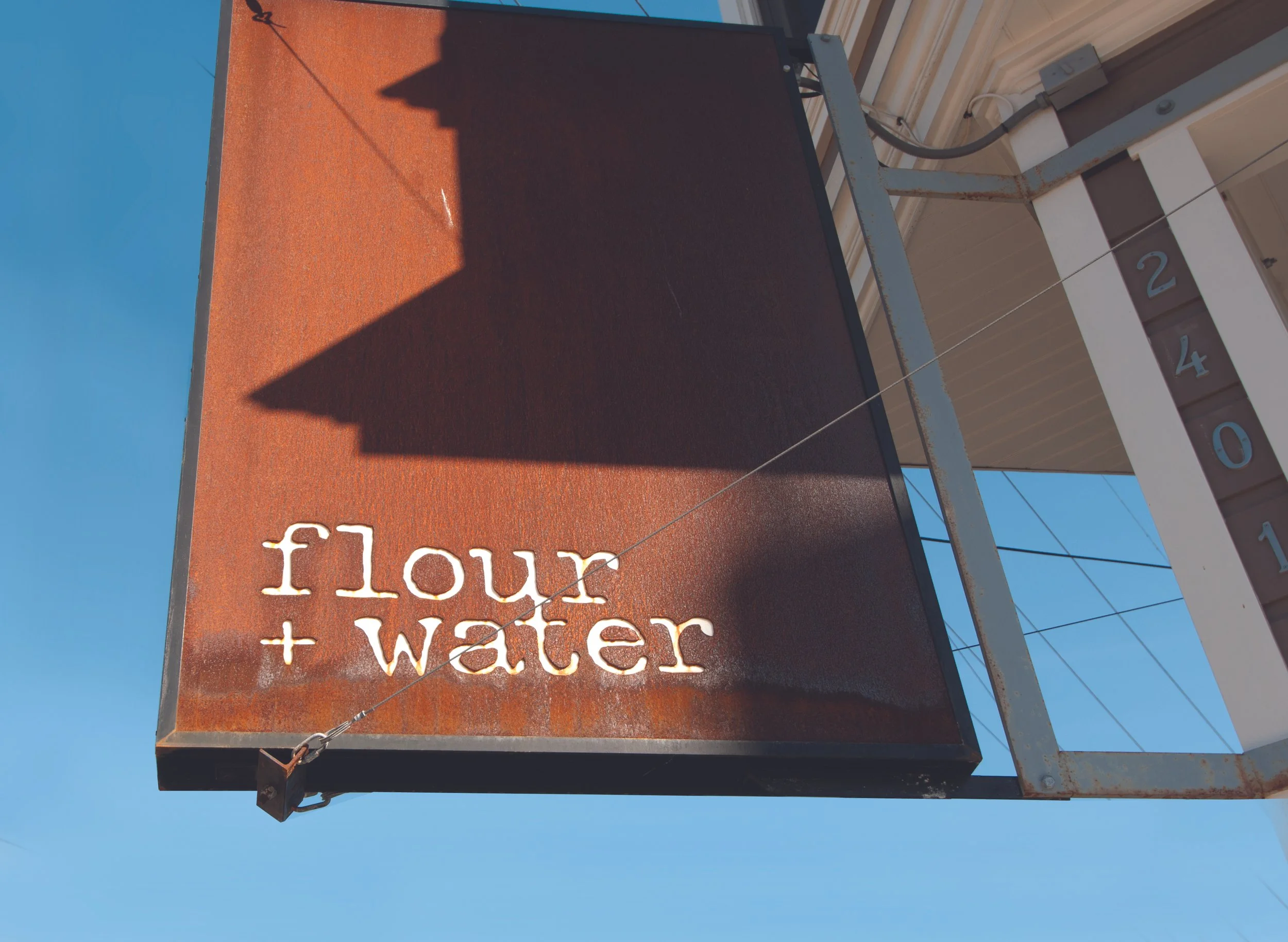 Rusty outdoor sign with white text that reads 'flour + water', mounted on a metal frame, with a clear blue sky in the background.
