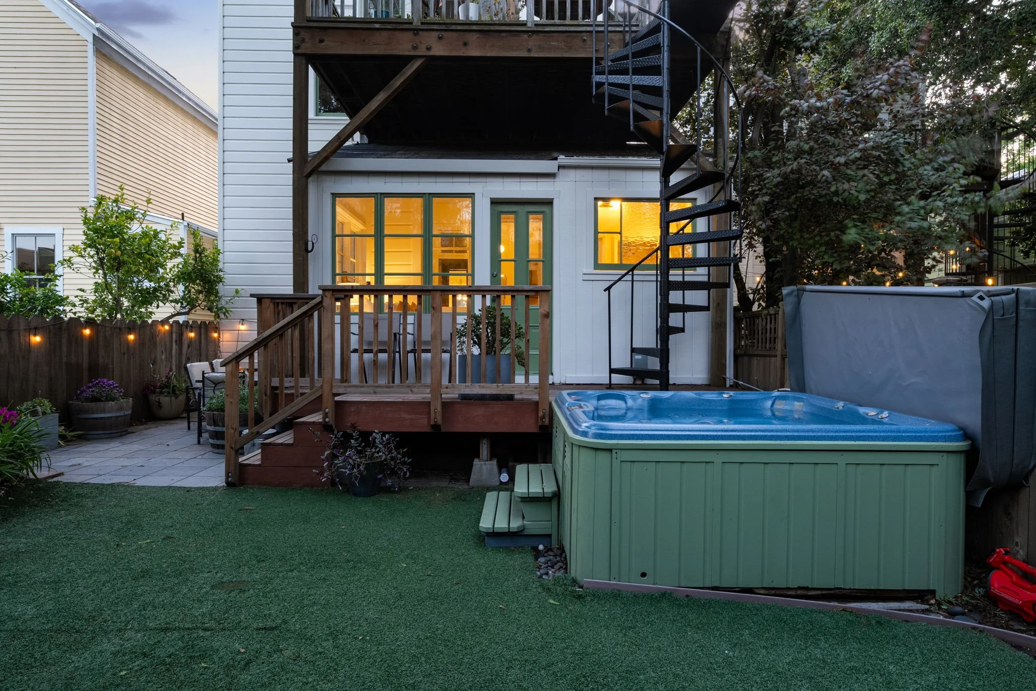 A backyard with a deck, spiral staircase, and a screened door leading inside, illuminated by warm interior lights, with a hot tub, potted plants, string lights, and trees surrounding the area.