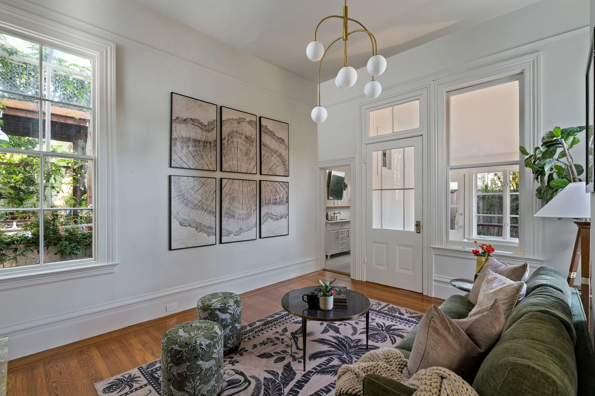 Living room with large windows, white walls, and wooden floors. Decor includes a modern chandelier with white globes, a framed woodgrain artwork, a green sofa with pillows, a black coffee table with a potted plant and books, a patterned rug, small gr