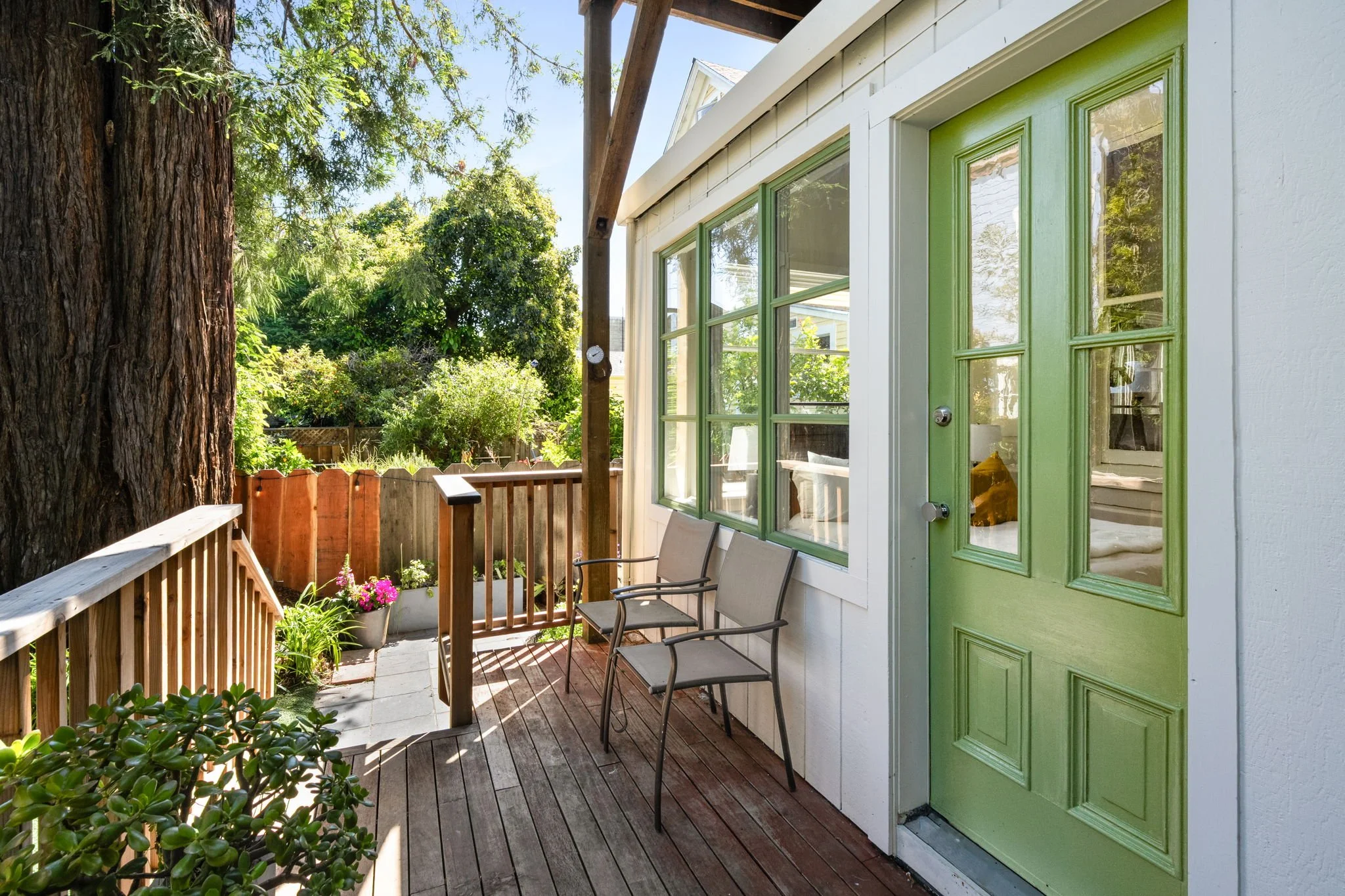 A small outdoor patio with a wooden deck, two patio chairs, potted plants and flowers, a wooden fence, large trees, and a white house with a green door and window trim. The scene is sunny with clear skies.