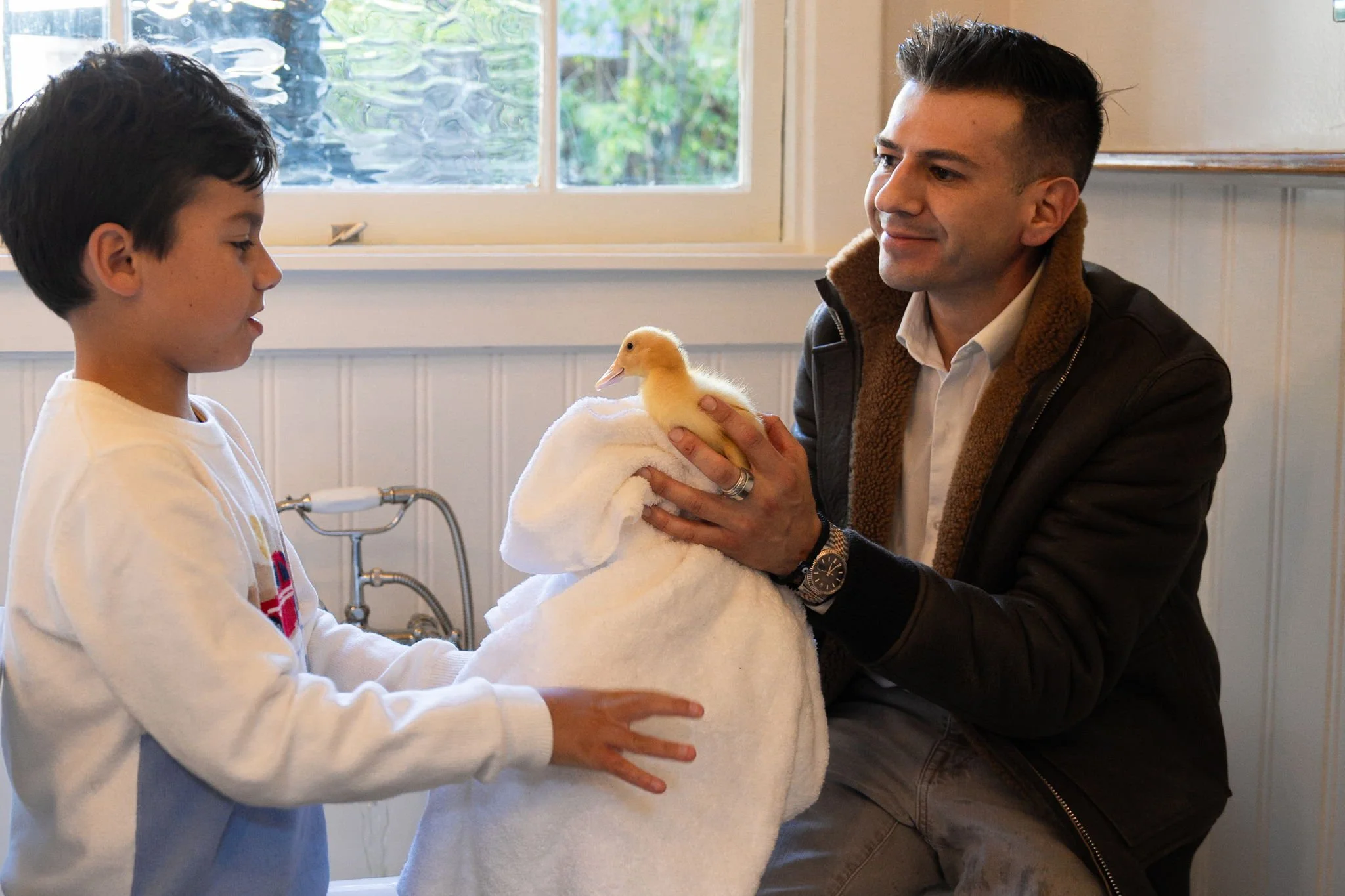A young boy and a man holding a yellow duckling wrapped in a towel in a room with a window