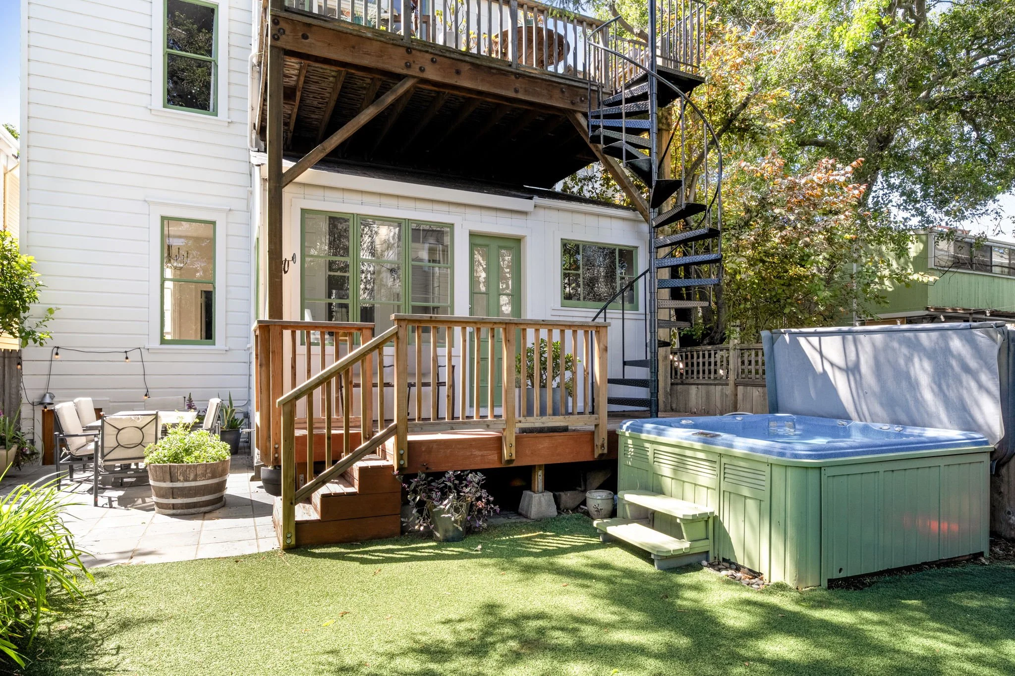 Backyard with patio furniture, potted plants, lawn, and a wooden deck with spiral staircase leading to an upper level, surrounded by trees and neighboring houses.