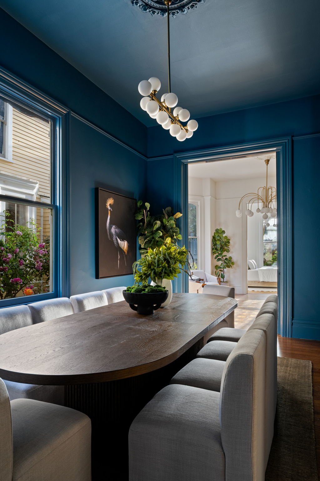 A dining room with blue walls, white upholstered chairs around a wooden table, and a dark blue wall with a large framed photo of a bird. There is a large pot with green plants on the table, and windows letting in natural light.