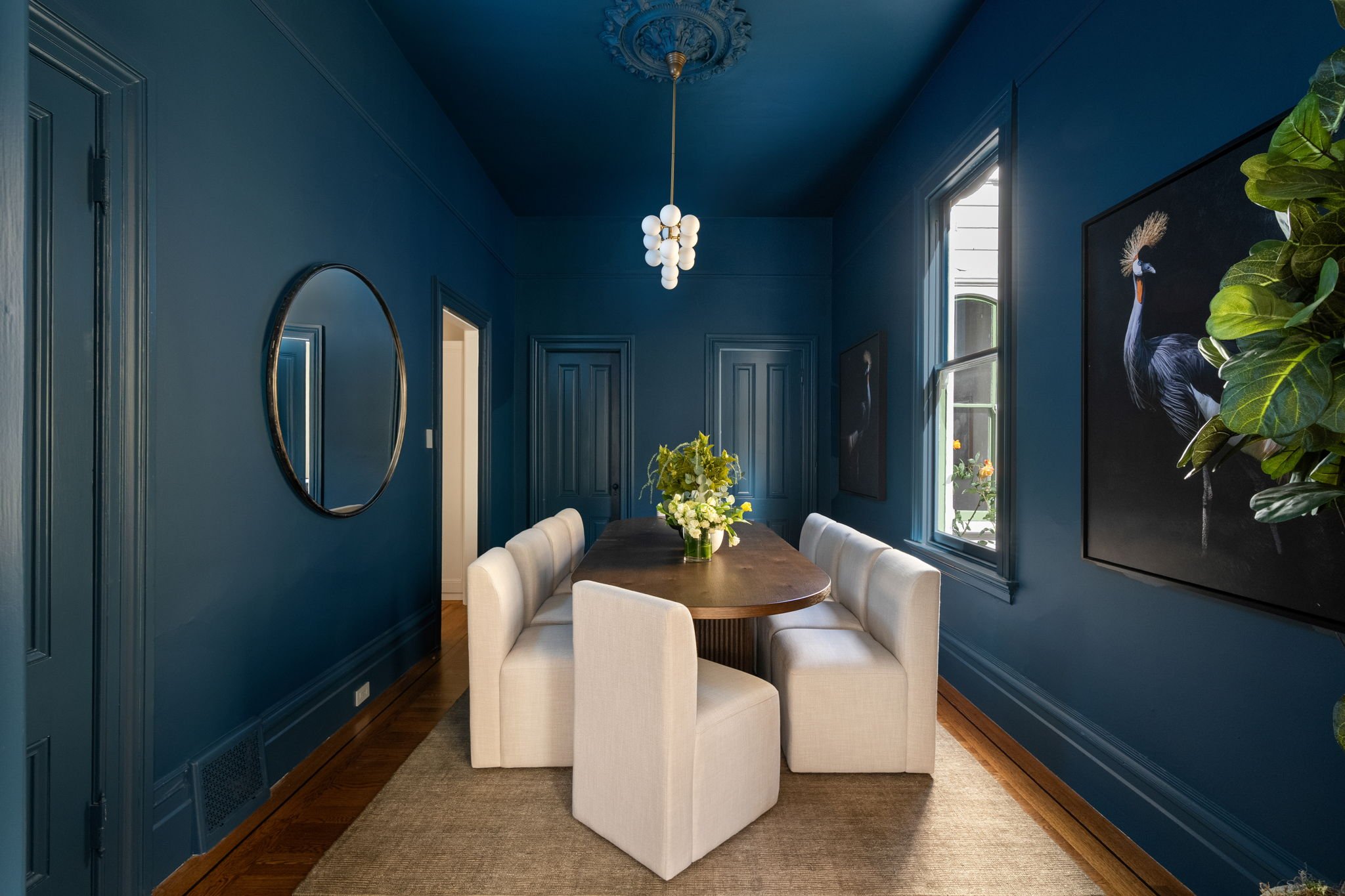 A dining room with dark blue walls, a round wooden table, white upholstered chairs, a glass vase with white flowers, a window, and framed artwork of a peacock on the wall.